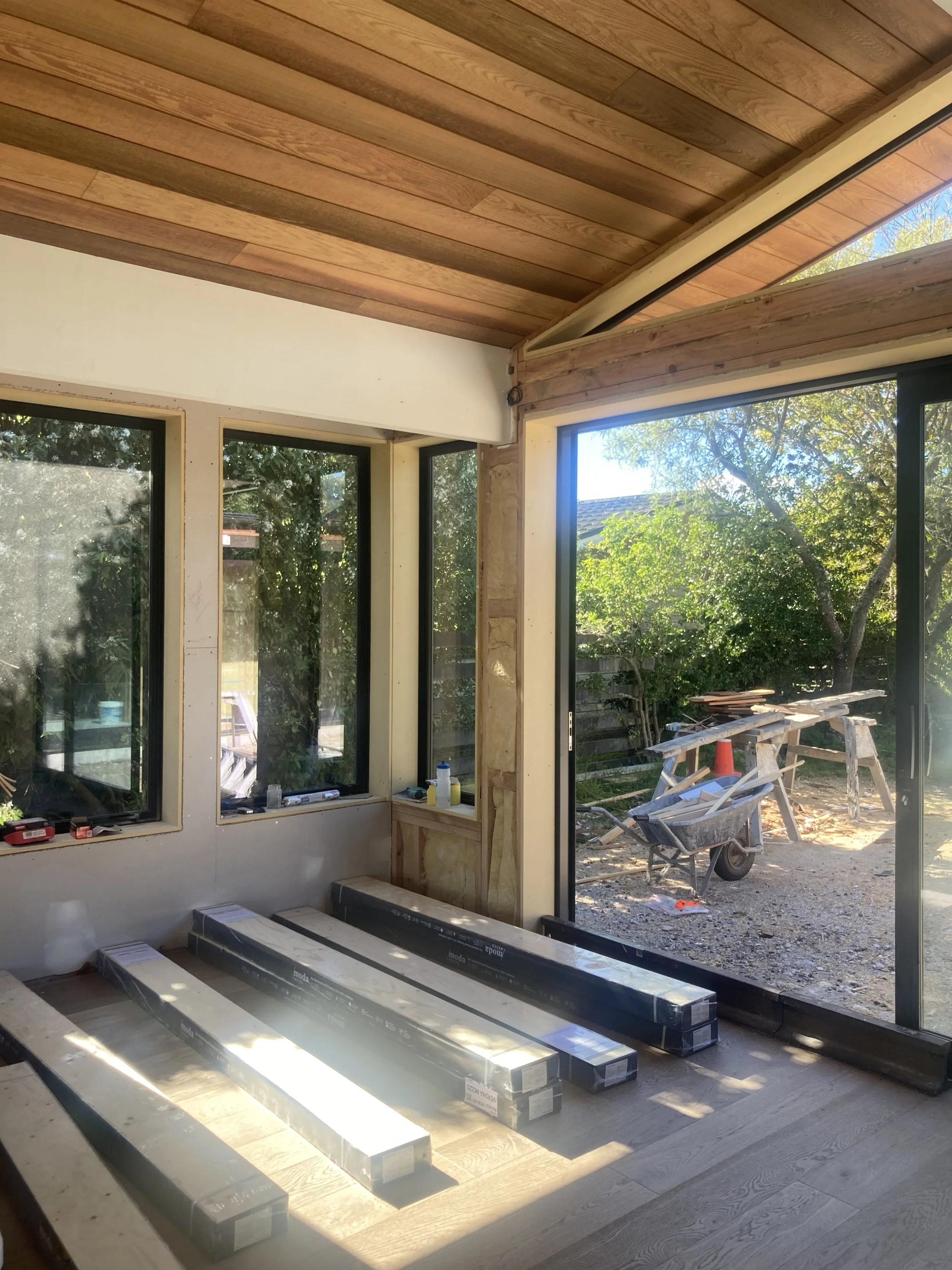 Interior view of a room under construction with large windows and sliding glass door, showing wooden ceiling, floor, and construction materials including long boxed pieces of flooring or trim.