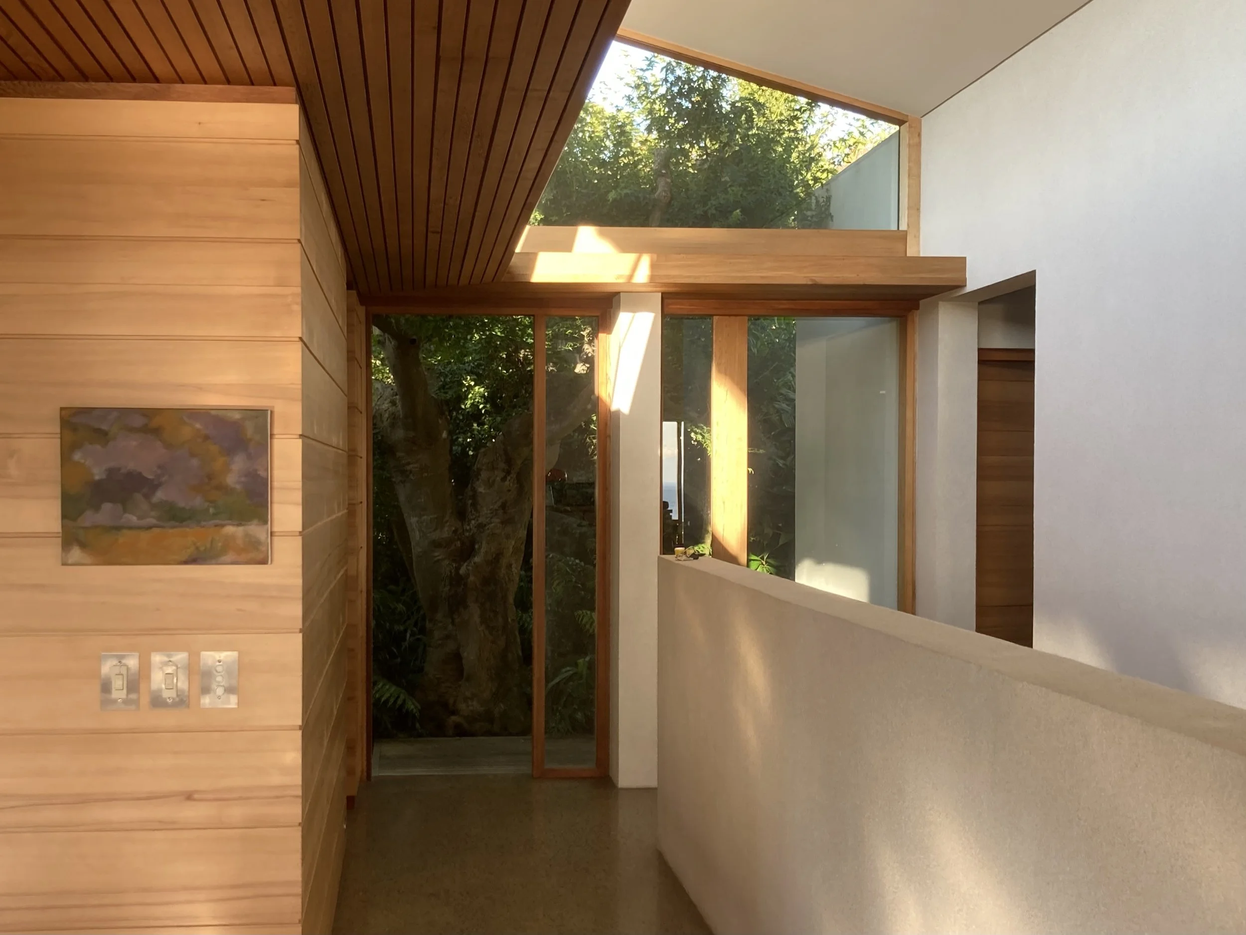Interior of a modern house with wooden paneling, large glass windows showing trees outside, and a white wall with a painting and light switches.