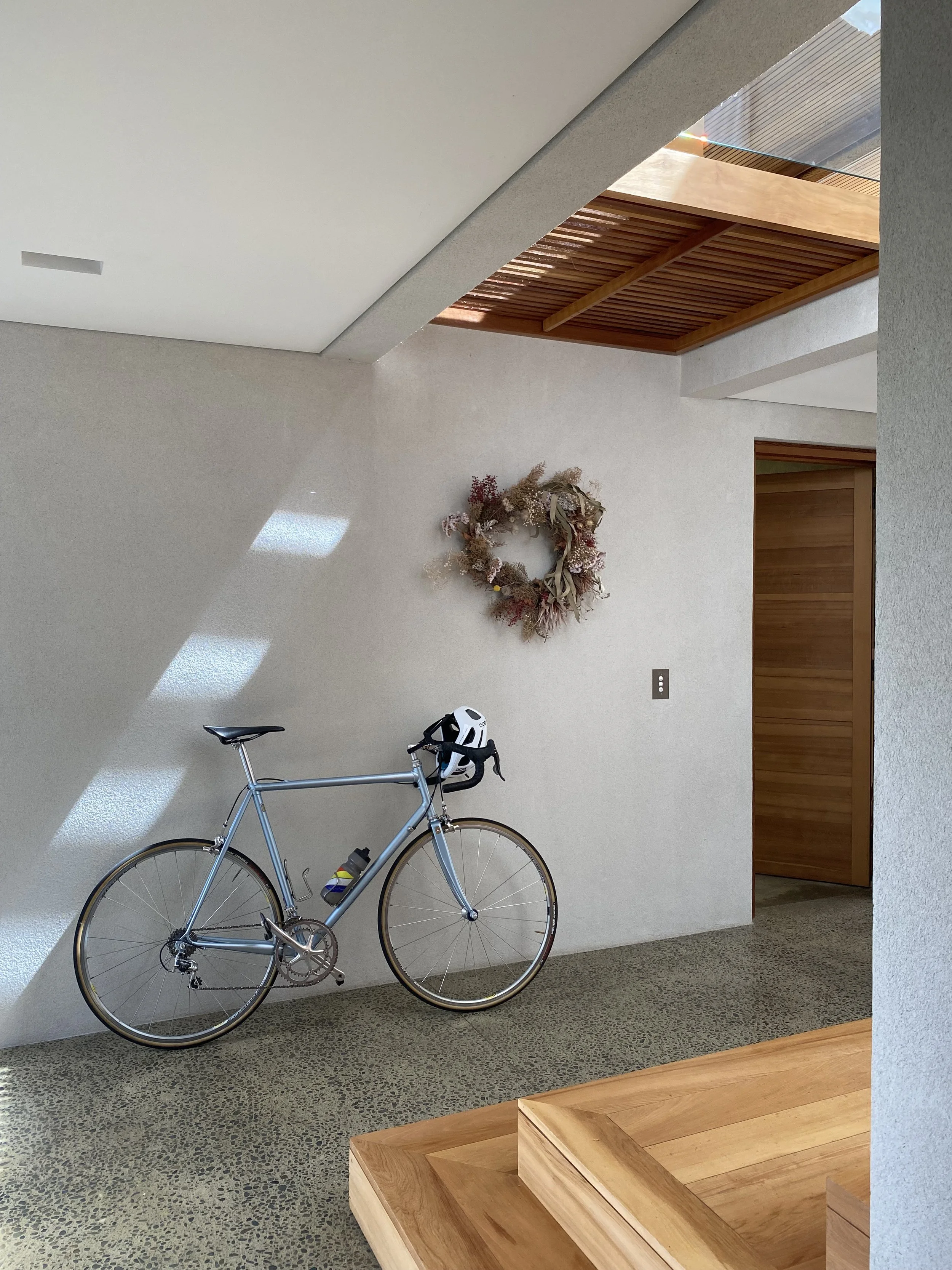 Modern interior with a silver bicycle leaning against a light-colored wall, a decorative wreath on the wall, and a wooden feature wall with a ceiling cutout, sunlight creating shadows on the wall, and wooden flooring.