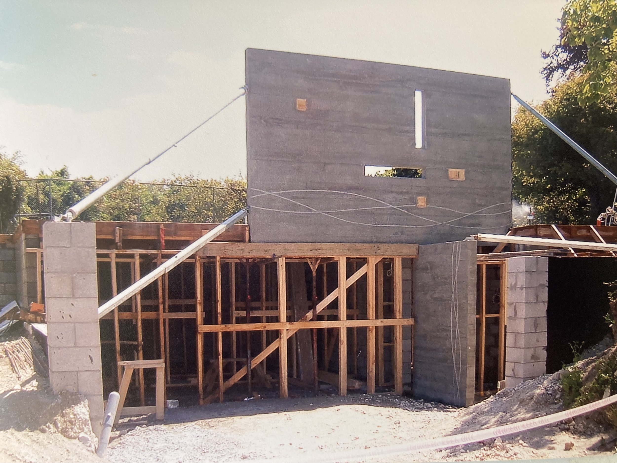 Construction site with concrete block walls, wooden framing, and a large gray concrete wall partially installed, surrounded by trees and construction equipment.