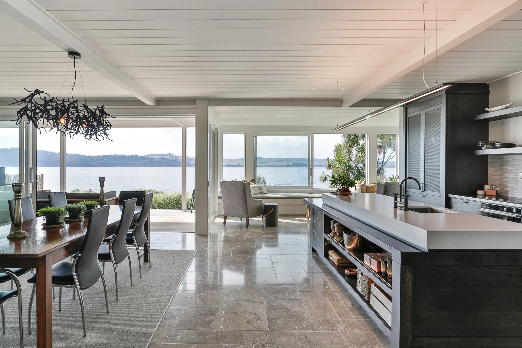 Open-concept kitchen and dining area with large windows overlooking a lake, with a wooden dining table and gray chairs, and a kitchen island with black cabinets and a white countertop.