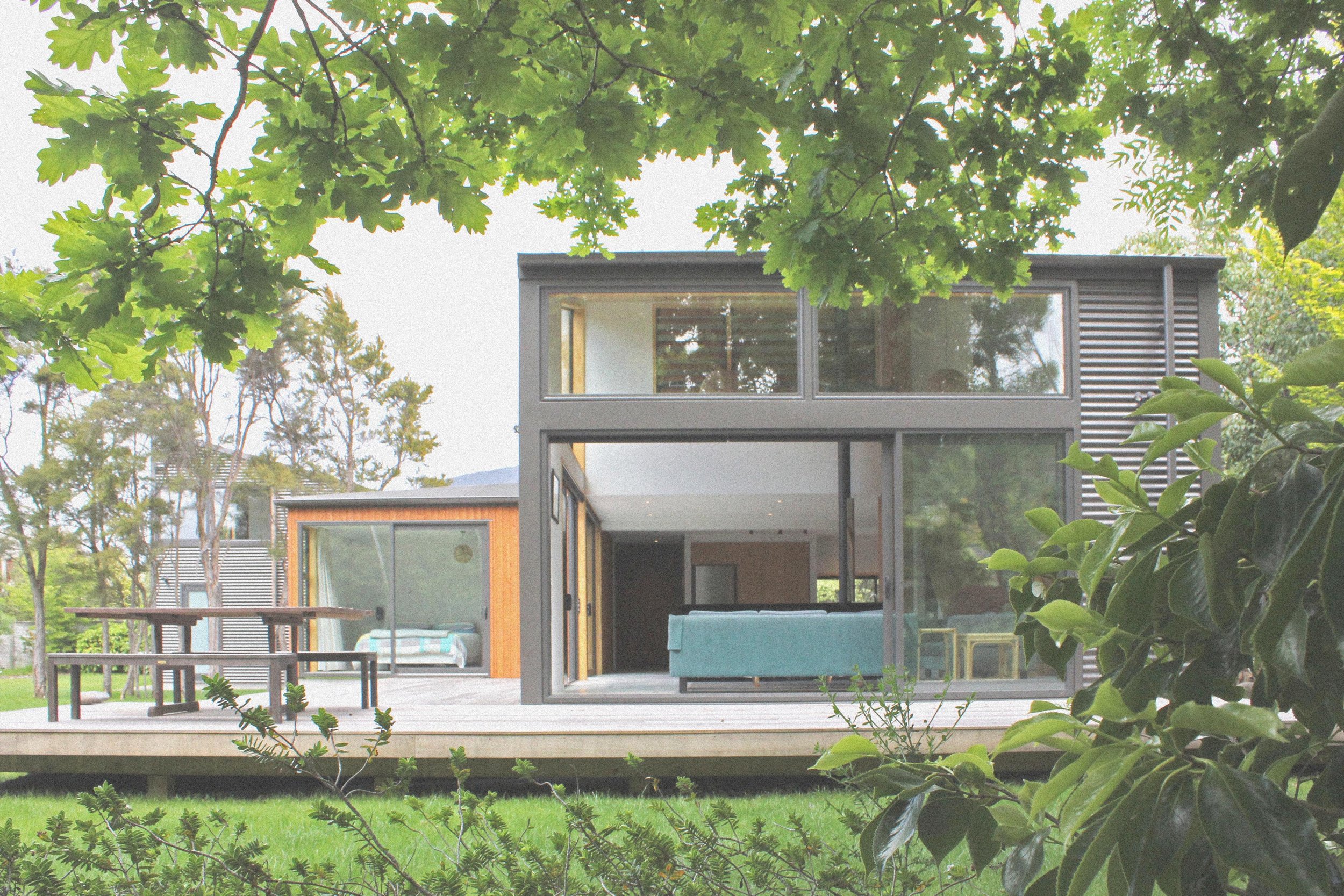 Modern house with large glass windows, surrounded by greenery and trees, with outdoor deck and outdoor furniture visible. Bach Design New Zealand. Bach Architect. Holiday Home Architect. Rural Architect. 