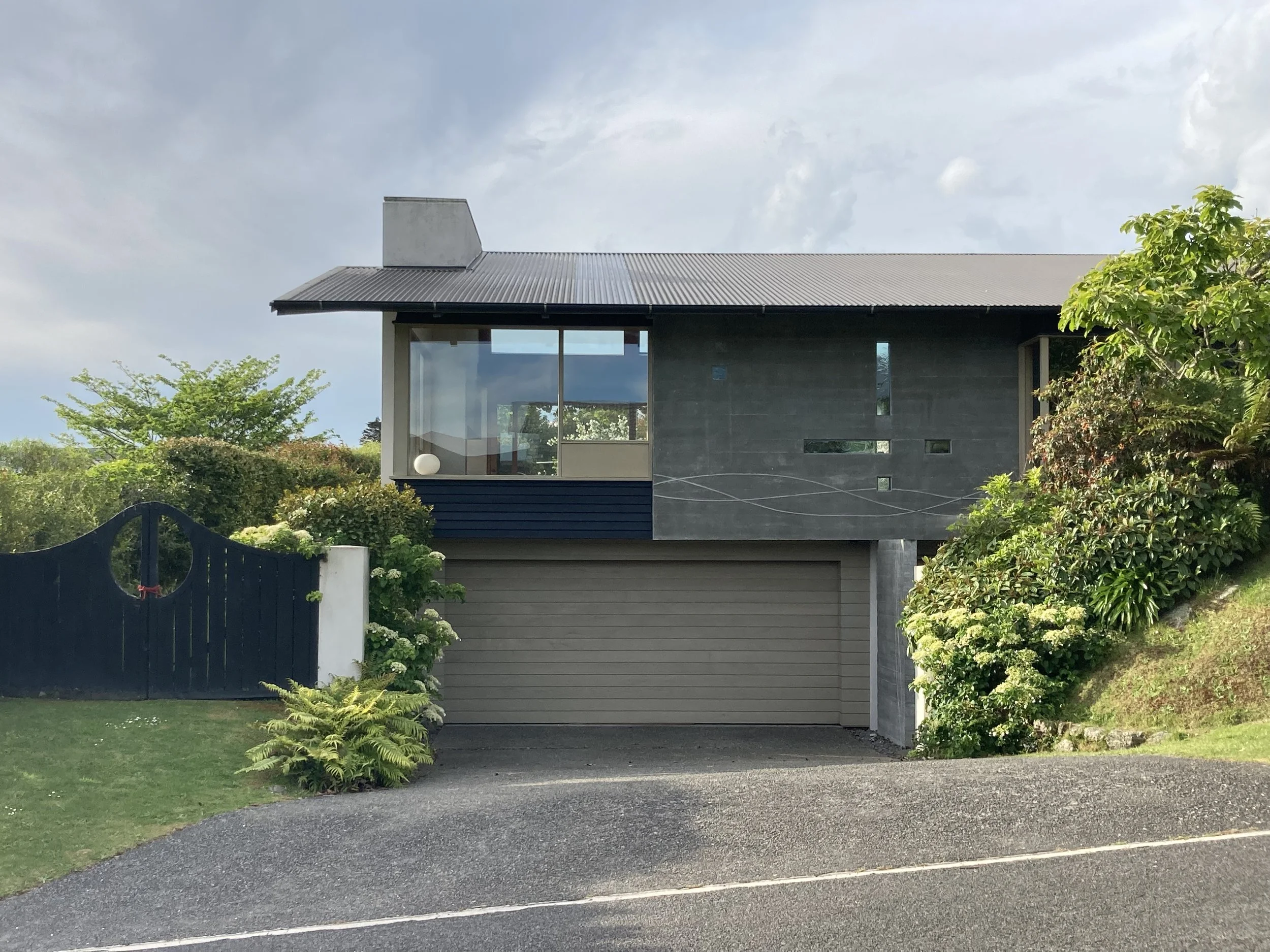 Modern two-story house with grey and black exterior, glass windows, a garage on the ground level, surrounded by greenery and a driveway in the foreground.