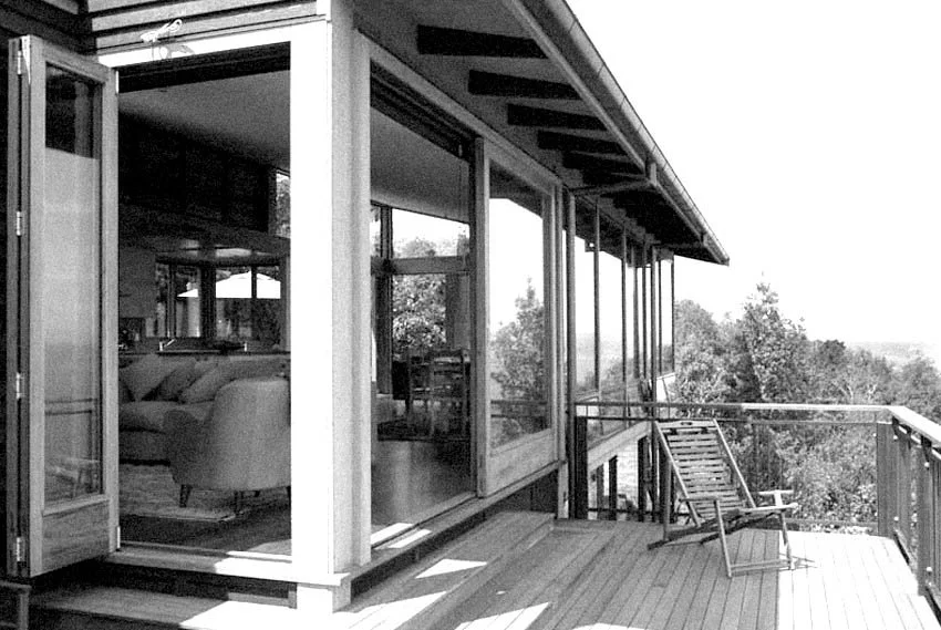 View of a house's outdoor deck with a wooden chair, overlooking a wooded landscape in black and white.