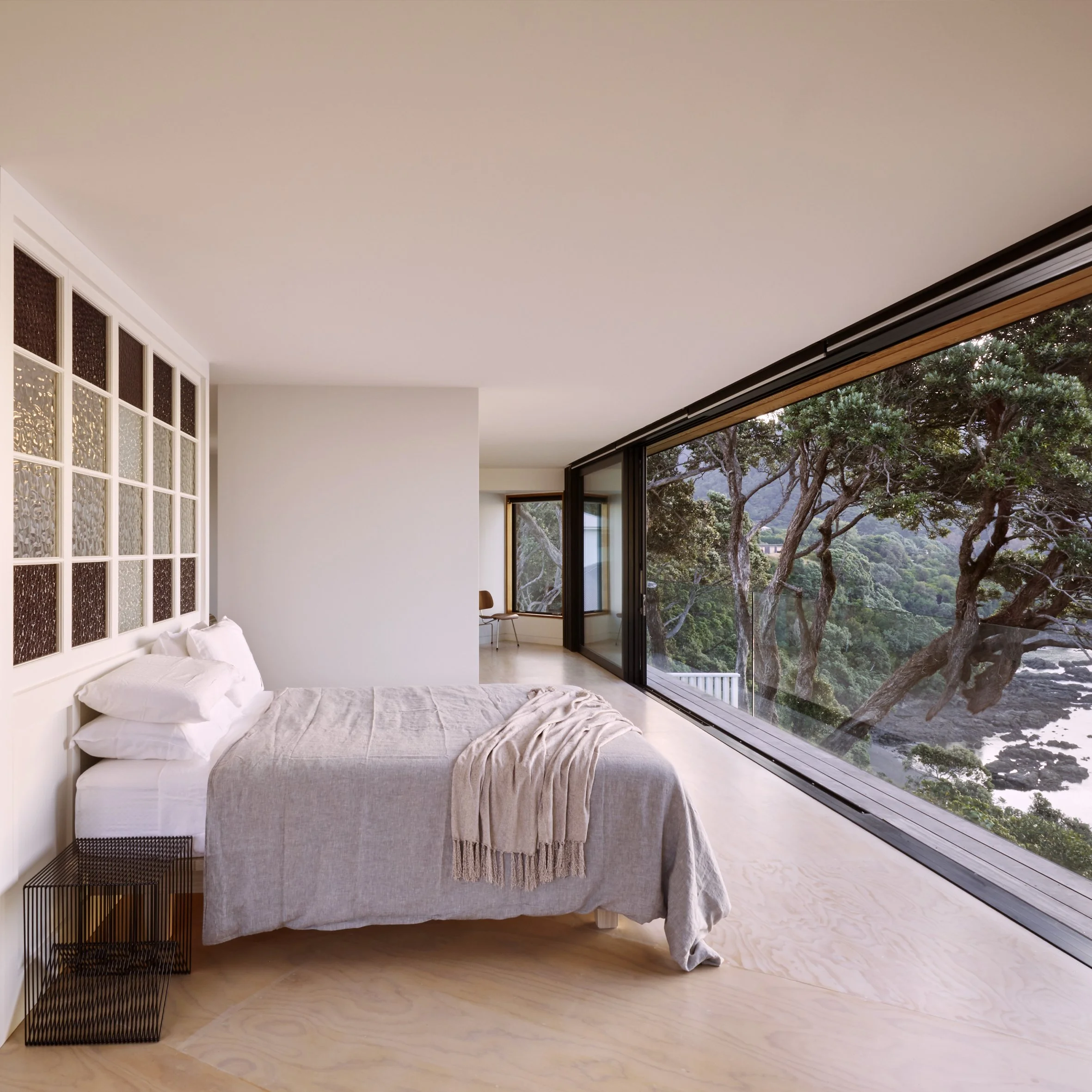 Modern bedroom with a large window showing a view of trees and water, white bed with gray blanket, corner chair, minimalist decor.