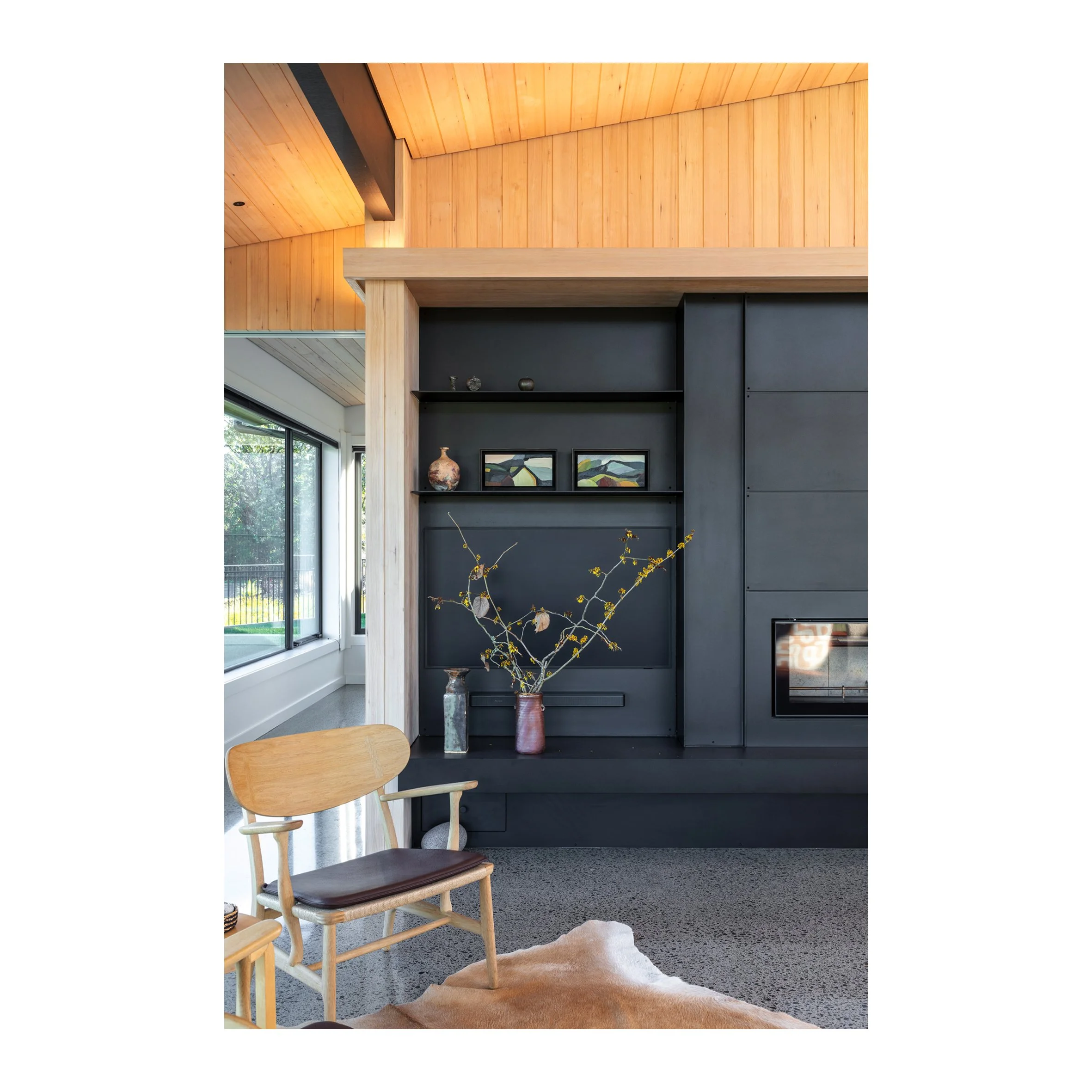 Interior living room with black built-in wall unit, decorative vases, framed artwork, large window with view of outdoors, wooden ceiling, beige chair, and a cowhide rug.