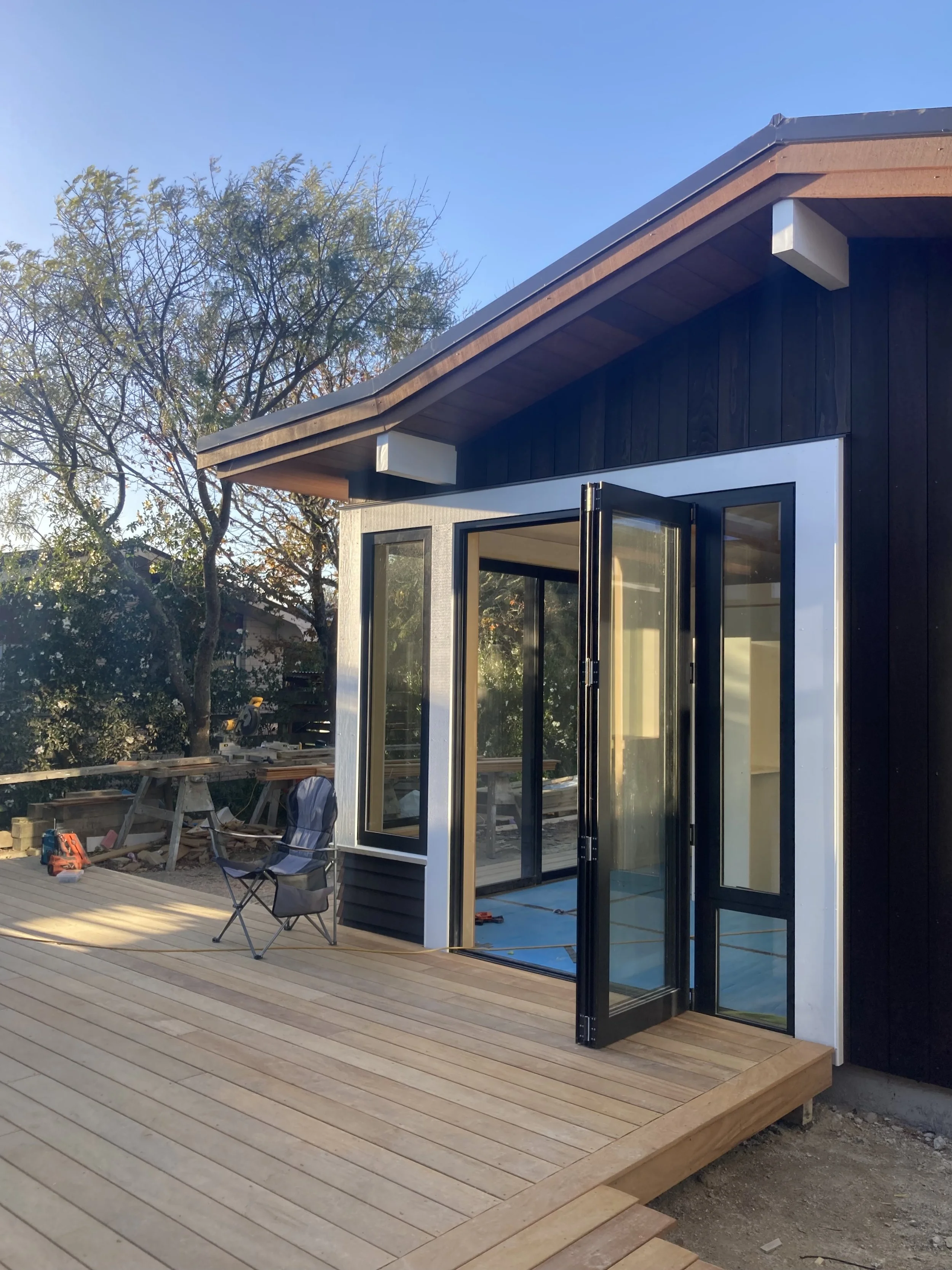 Construction of a house exterior with a black wooden siding, a sliding glass door, and a wooden deck. Construction tools and a chair are visible on the deck, with trees in the background.