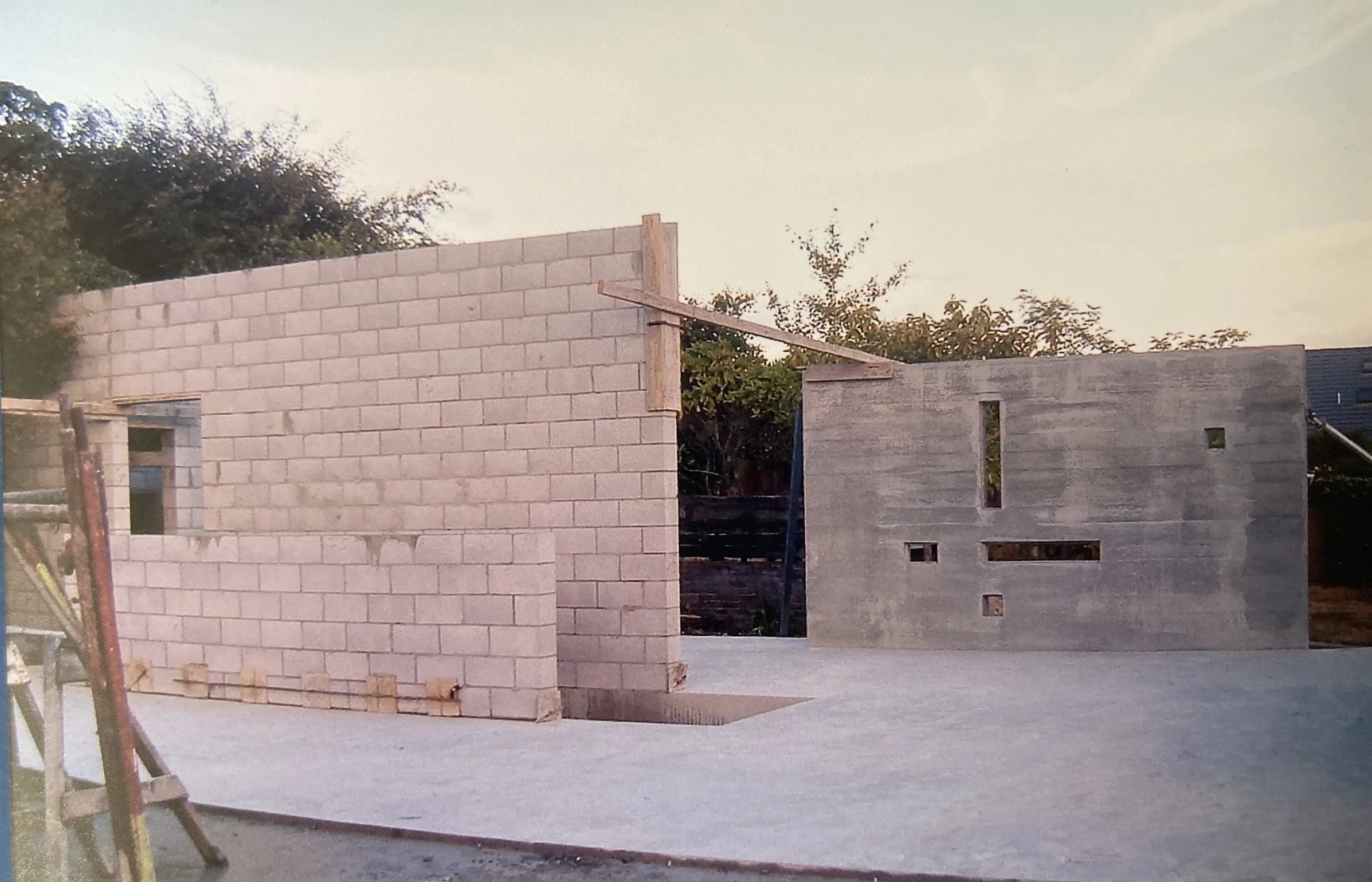 Construction site with partially built walls of concrete blocks and wood framing, overlooking trees and a clear sky.