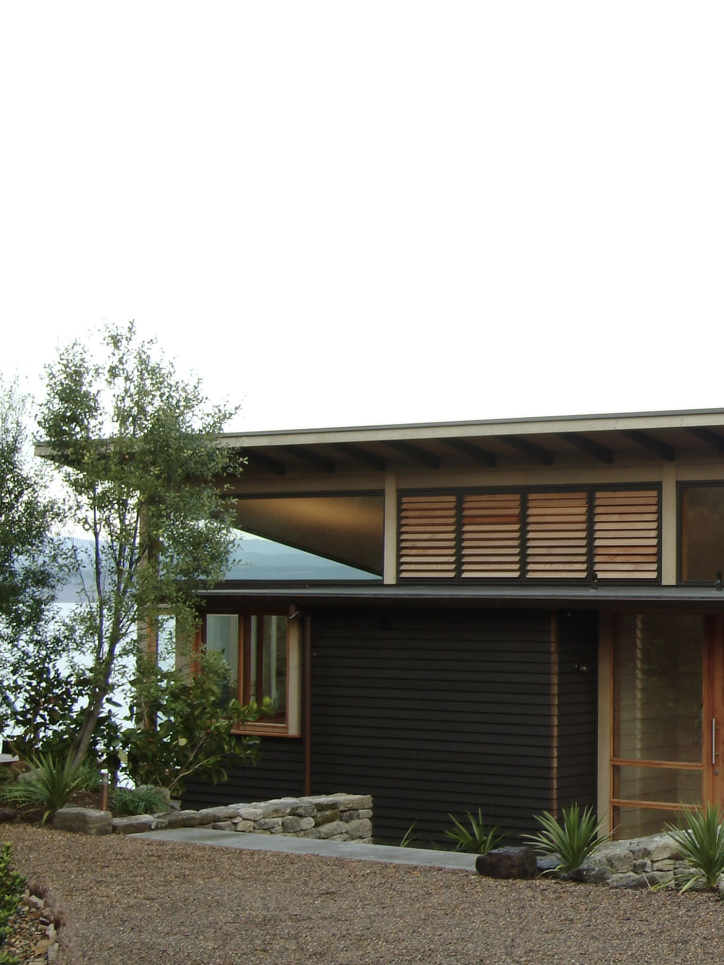 Modern house with wooden accents, black exterior, surrounded by gravel and plants, overlooking a body of water.
