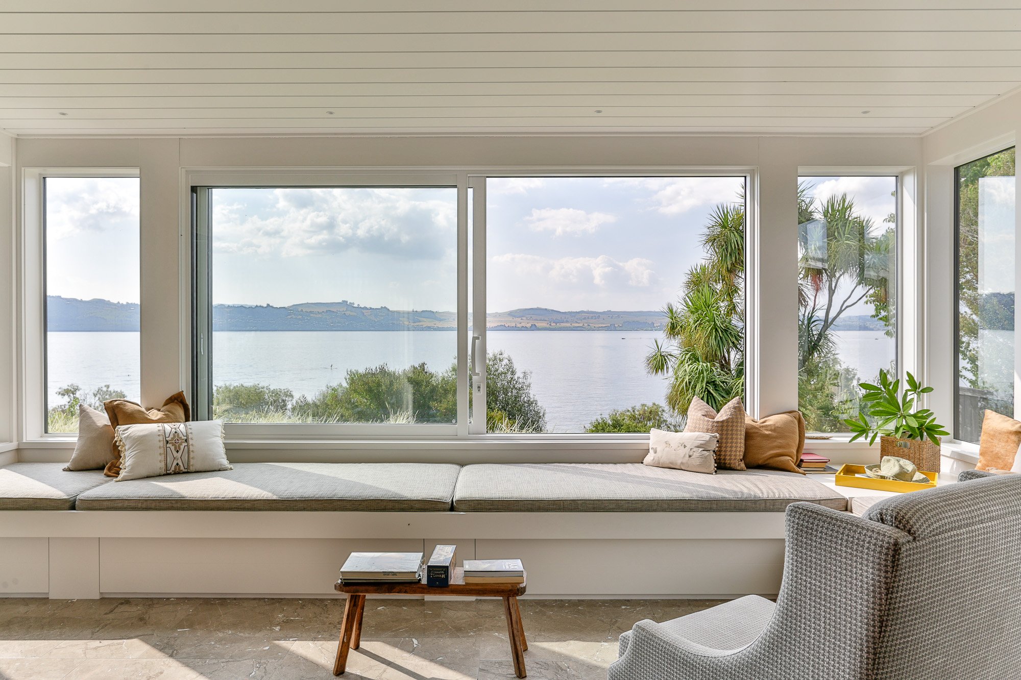 Living room with large windows overlooking water and landscape, cushioned window seat with pillows, small wooden coffee table with books, armchair, and indoor plants.