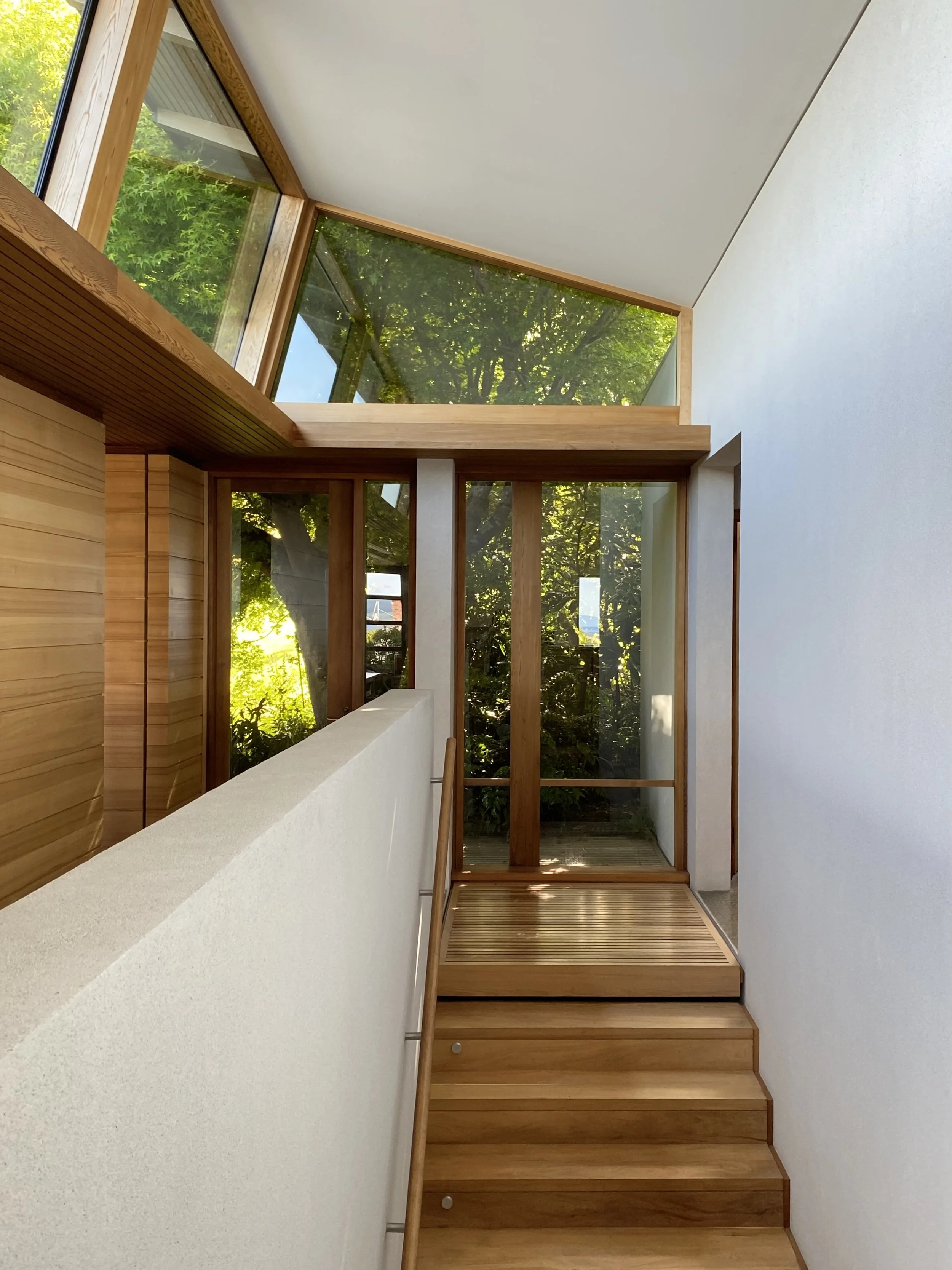 Interior view of a modern home with wooden stairs and large glass windows showing green foliage outside.