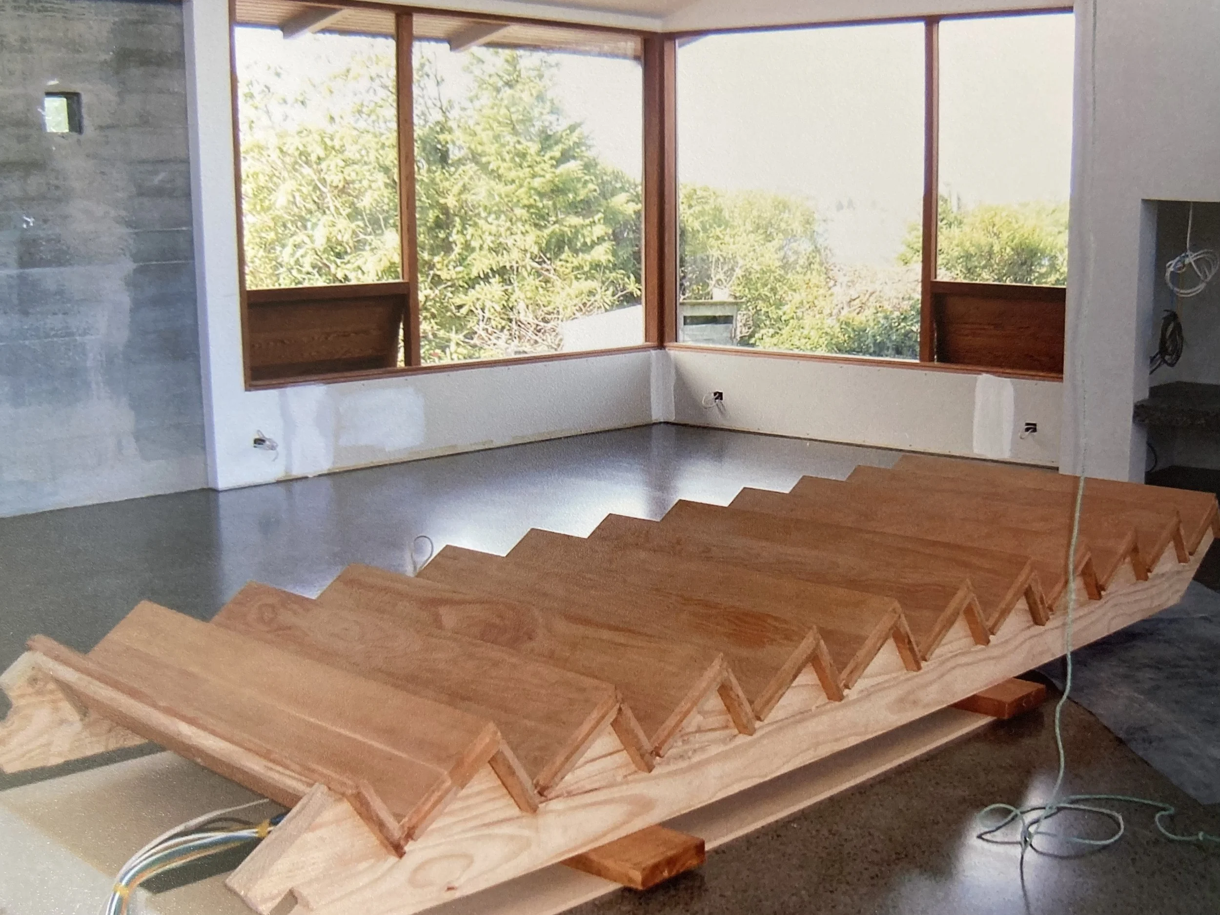 Interior of a house under construction with a large window overlooking green trees, a staircase frame made of wood, and unfinished walls with electrical wiring.
