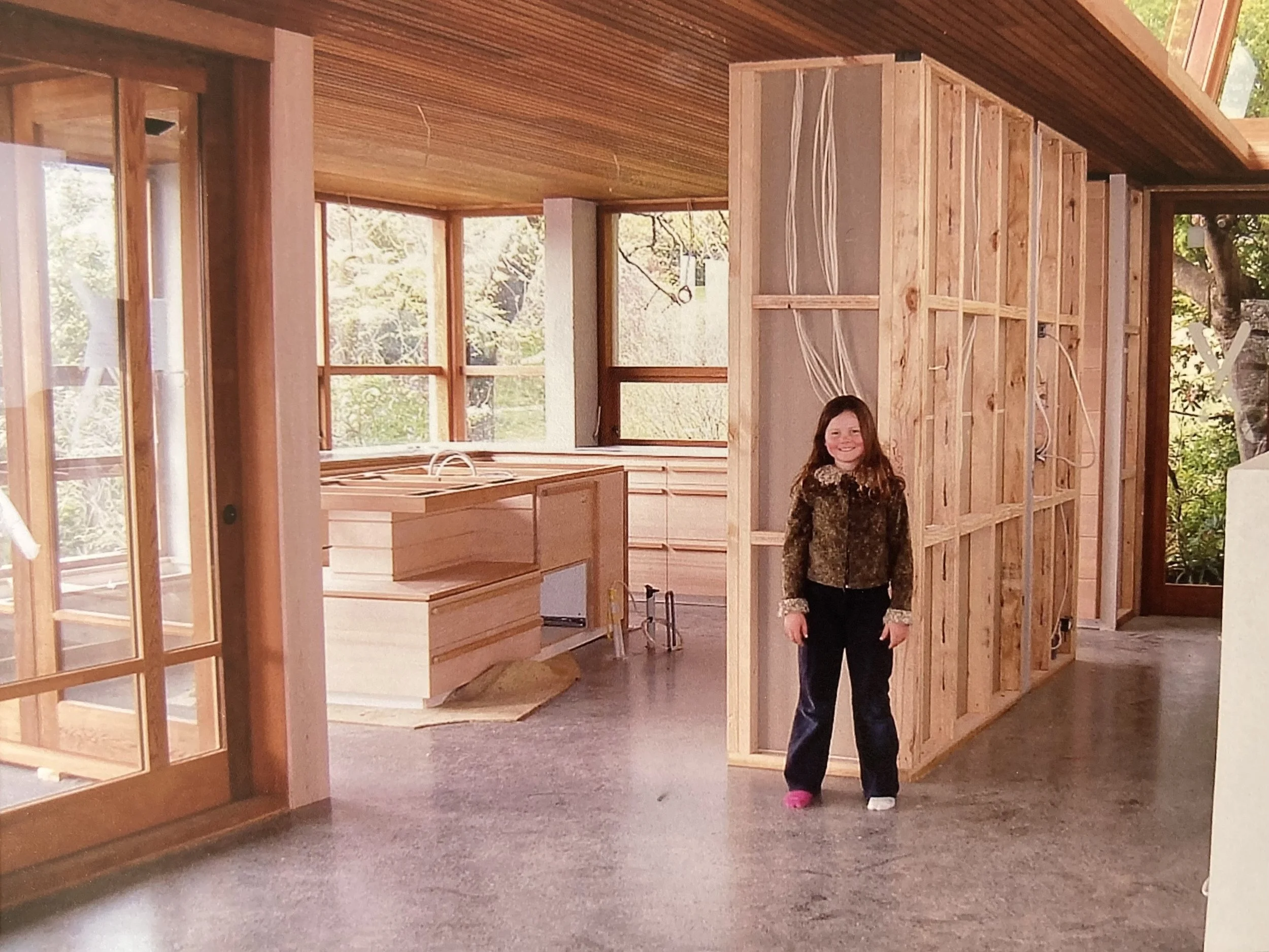 A young girl with long brown hair, wearing a brown and black patterned jacket and black pants, standing inside a house under construction, surrounded by visible wooden framing and large windows.