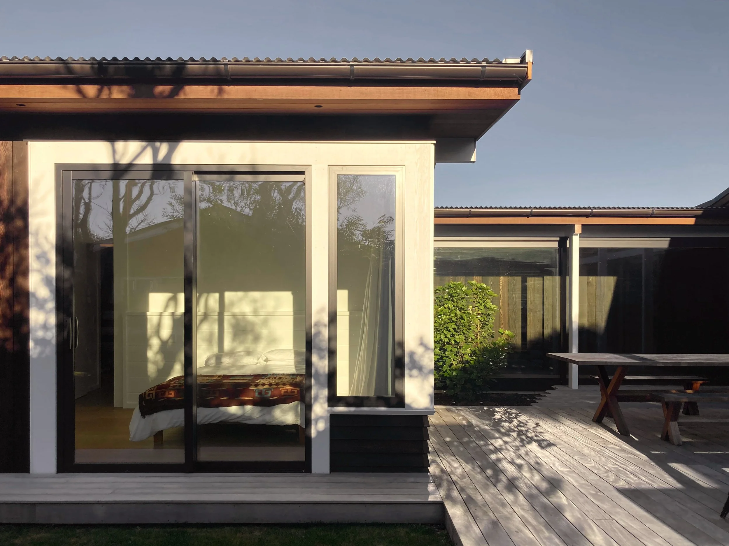Exterior of a modern house with large glass sliding doors, a wooden deck, and a picnic table, with shadows and greenery in the background.