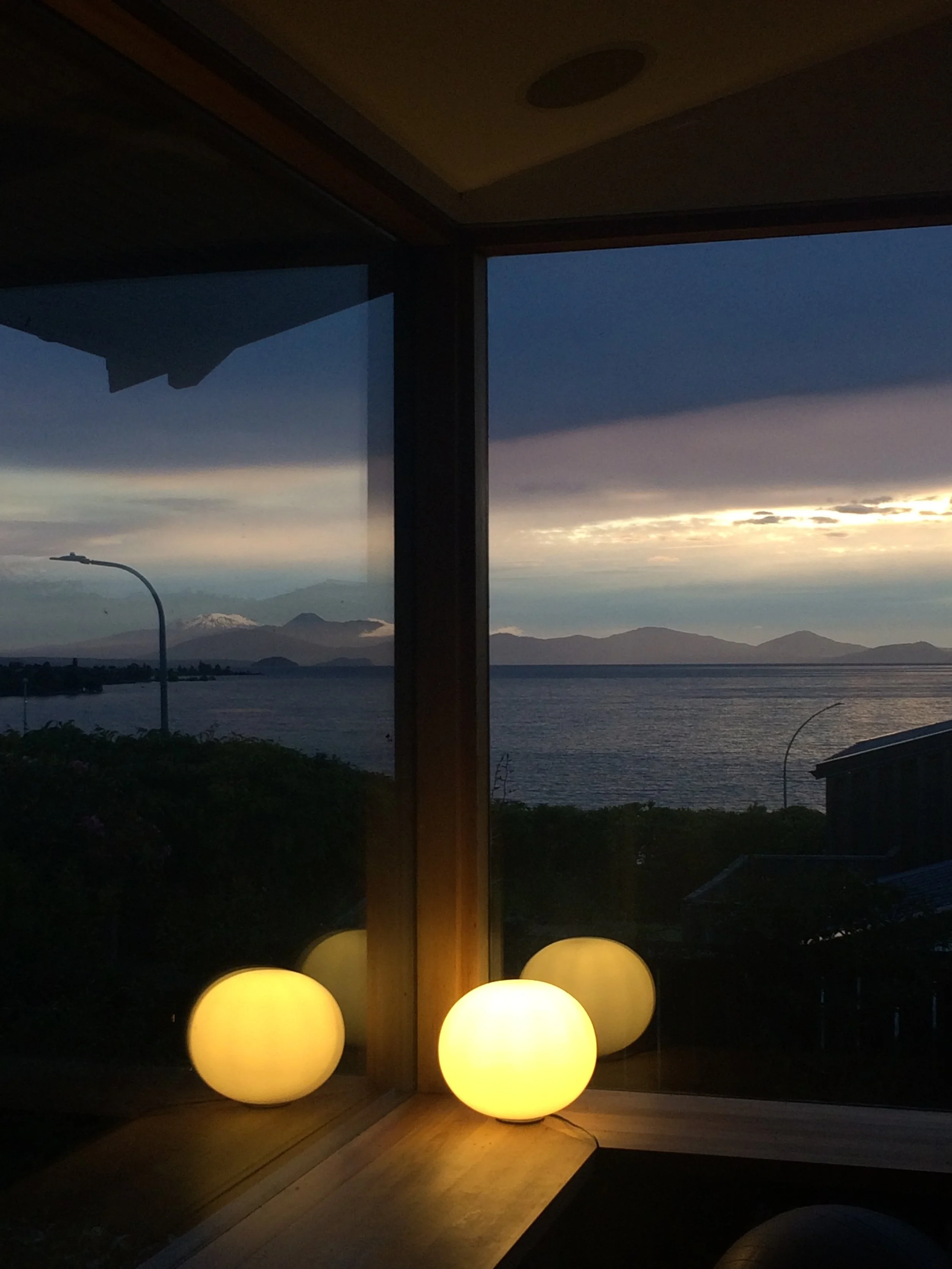 Interior view of a room with a large window showing a sunset over a body of water and mountains in the distance; three glowing round lamps on the windowsill