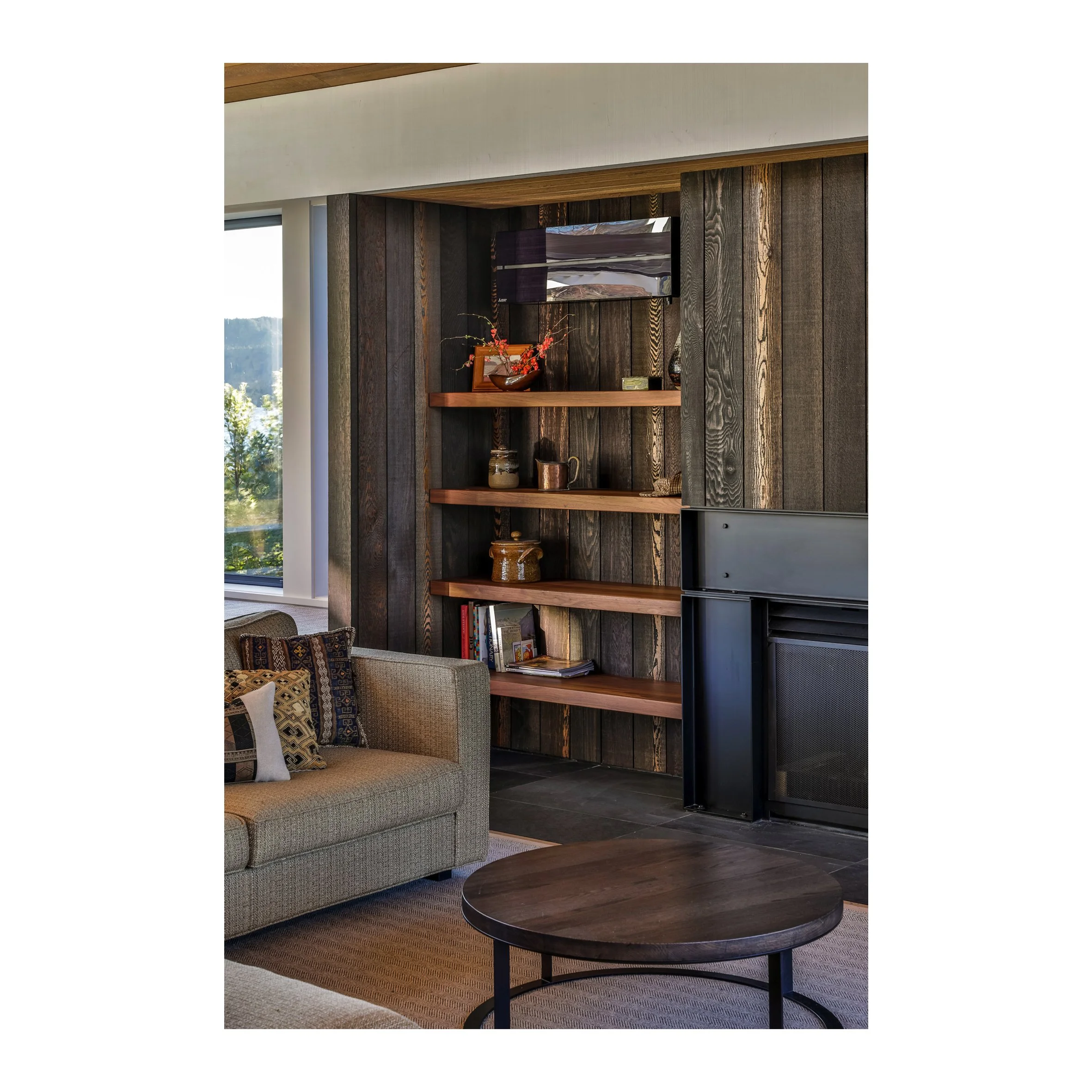 Modern living room with a gray sofa, patterned pillows, a round wooden coffee table, and a built-in wooden bookshelf with decorative items against a dark wood-paneled wall.