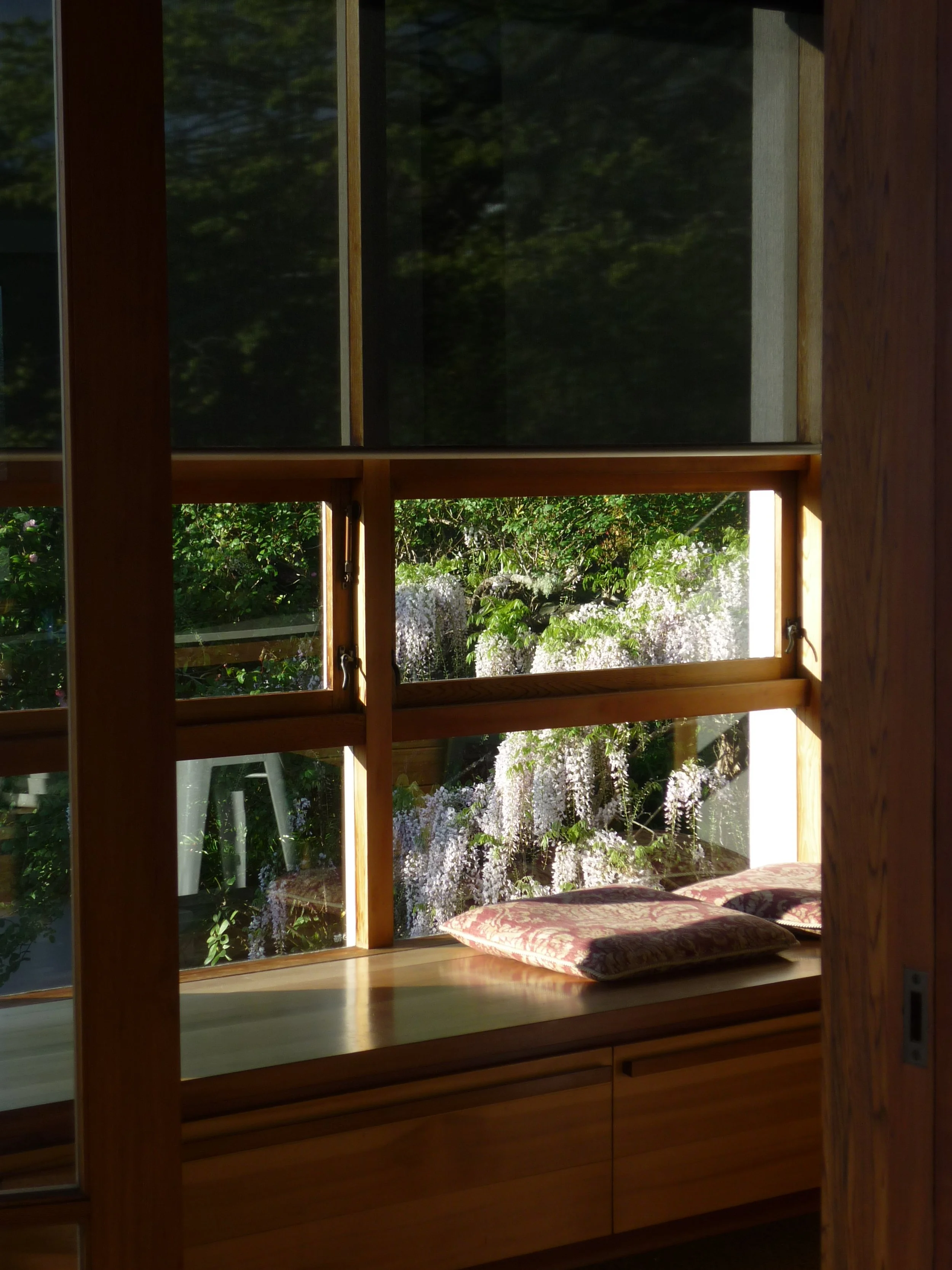 Inside view of a wooden window seat area with cushions, looking out onto a garden with white flowering plants.