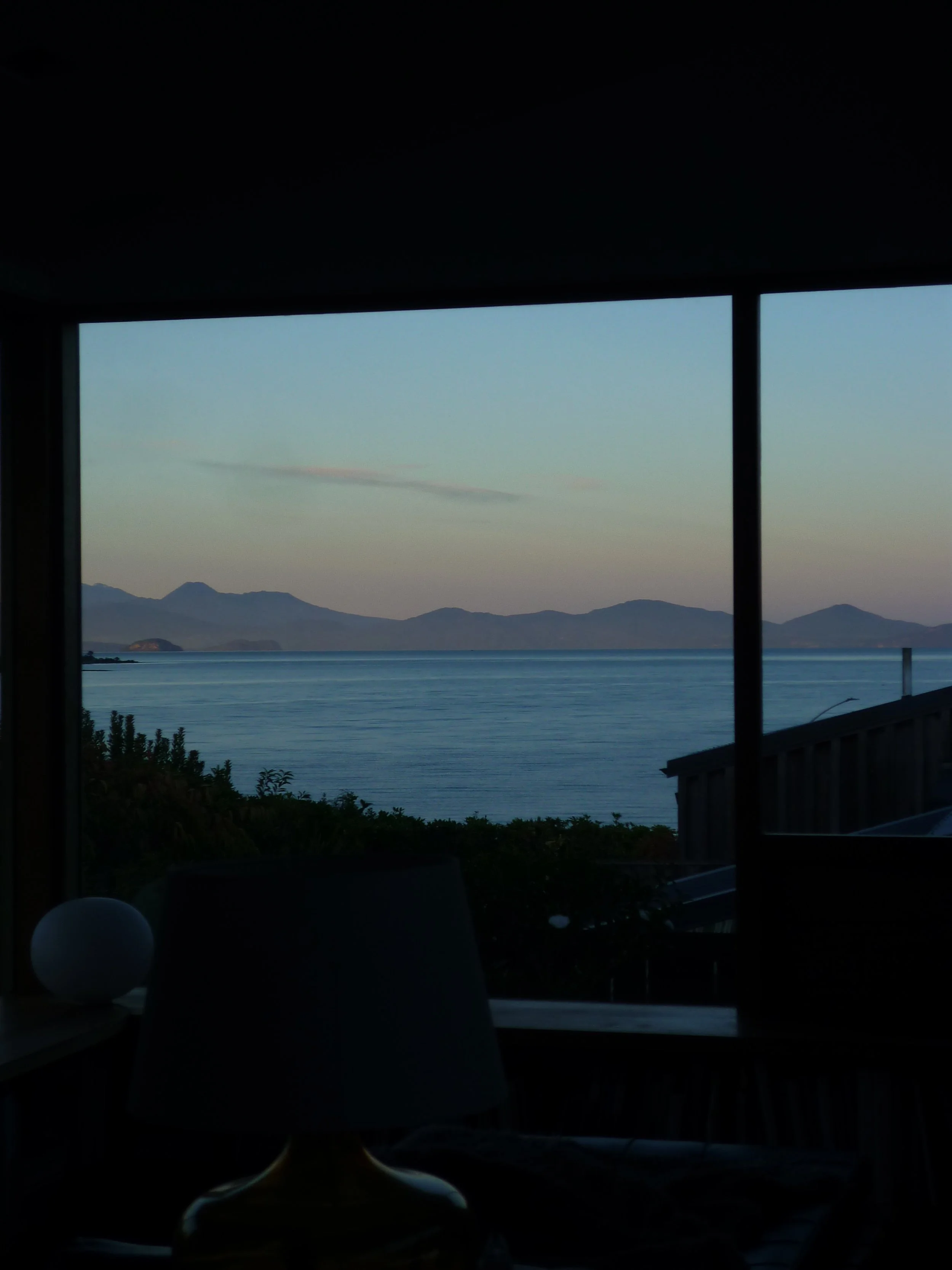 View of a calm body of water with distant mountains under a clear sky, seen through a window in a dimly lit room.