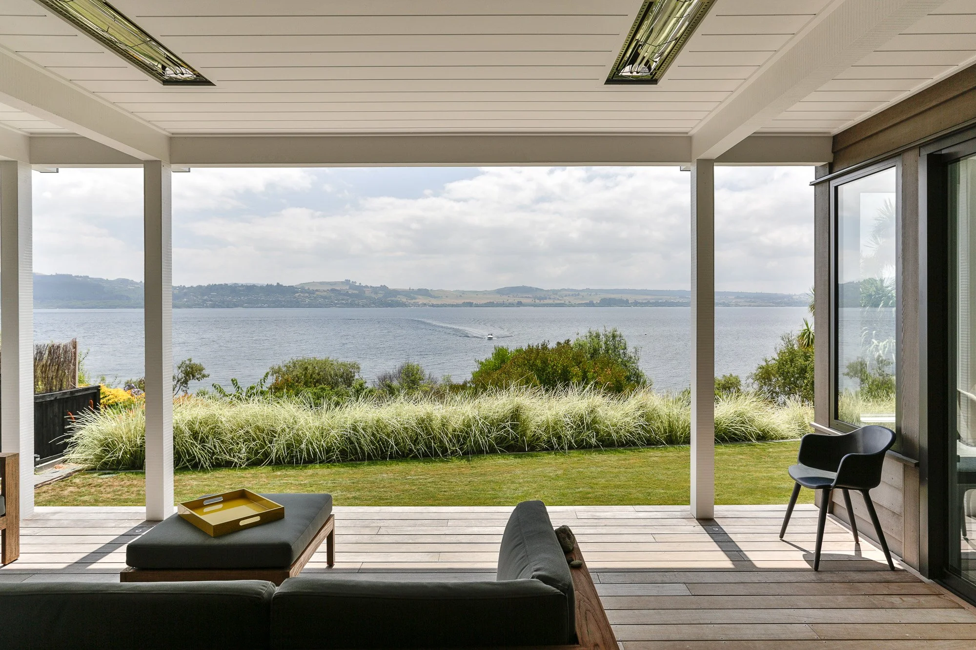 View of a house patio overlooking a lake with trees and hills in the background, outdoor furniture, and cloudy sky.
