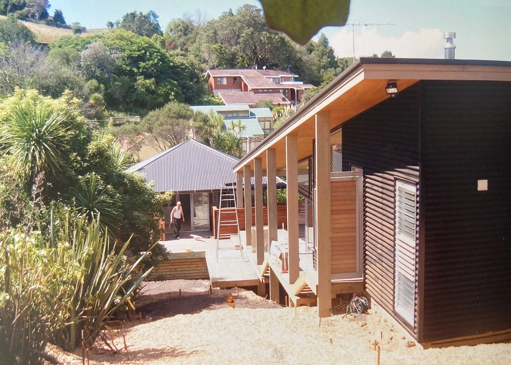 A person working on a wooden deck outside a house under construction, with a modern black and wood exterior, surrounded by lush green trees and other houses on a hillside. Bach Design New Zealand. Bach Architect. Holiday Home Architect.