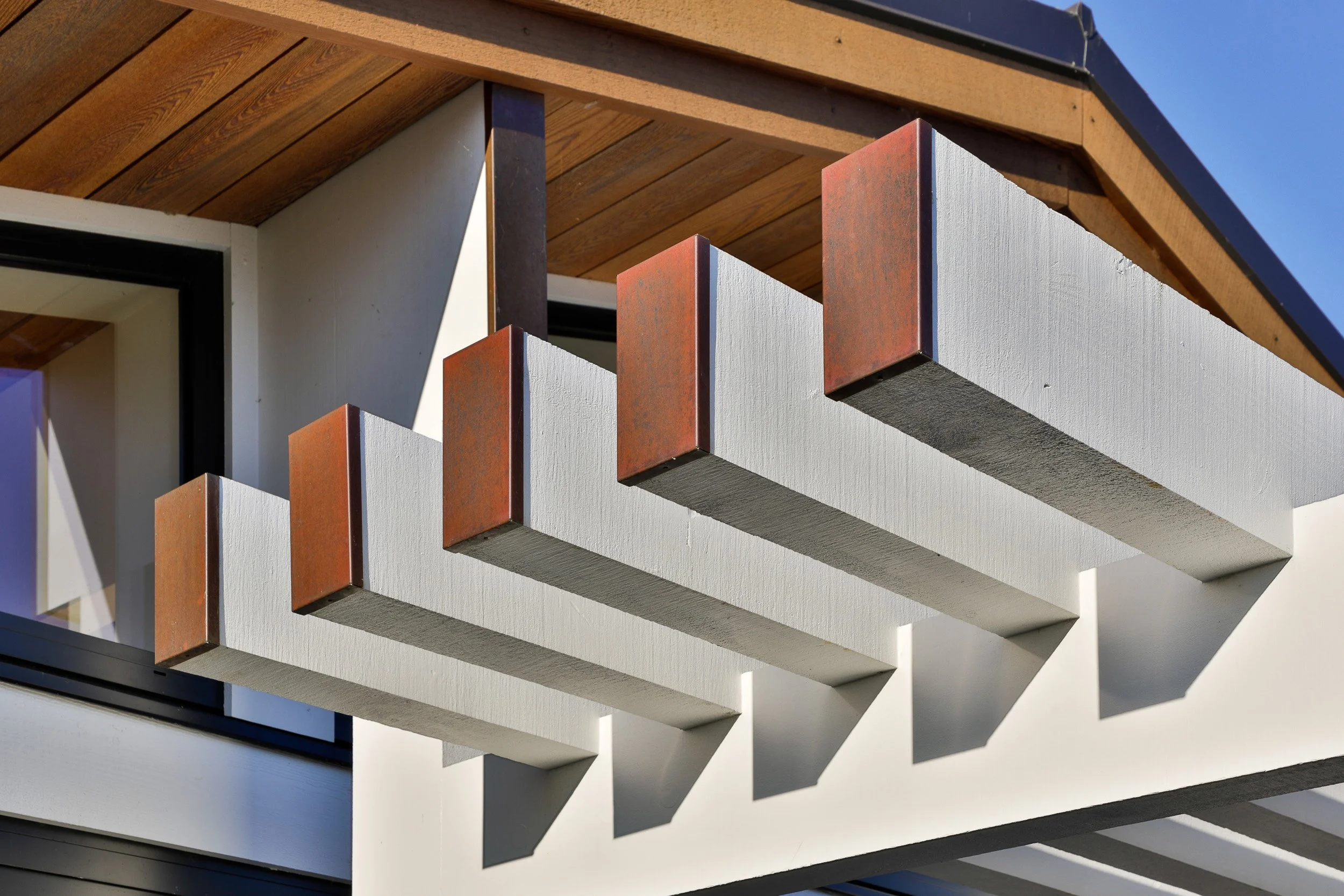Close-up of a modern house's exterior balcony with white wooden beams, rust-colored metal accents, and a wooden roof, casting shadows in sunlight.