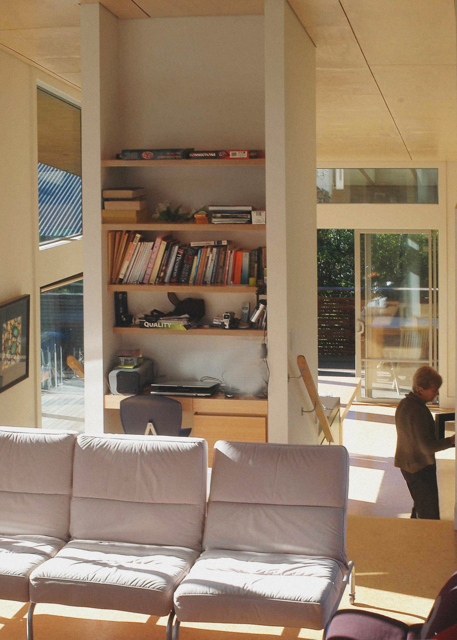 Interior of a home with sunlight streaming through large windows, a bookshelf with books and items, beige couch, and a child playing piano. Bach Design New Zealand. Bach Architect. Holiday Home Architect. Rural Architect. 