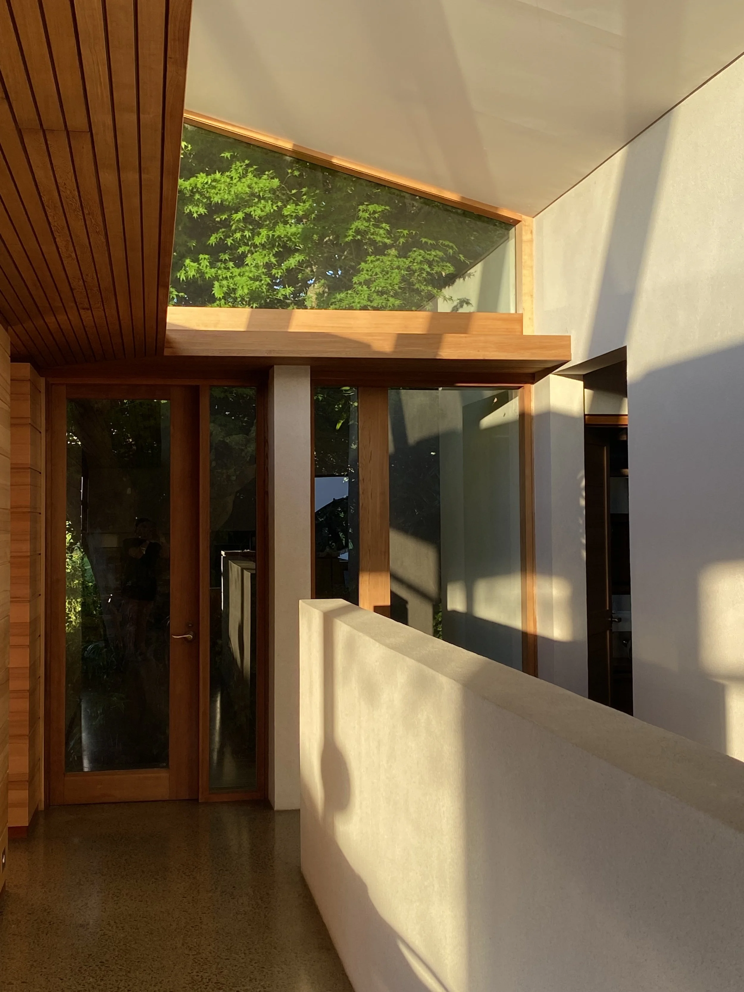 Interior view of a modern house entryway with wooden framing, glass door, and a large window letting in natural sunlight, with greenery visible outside.