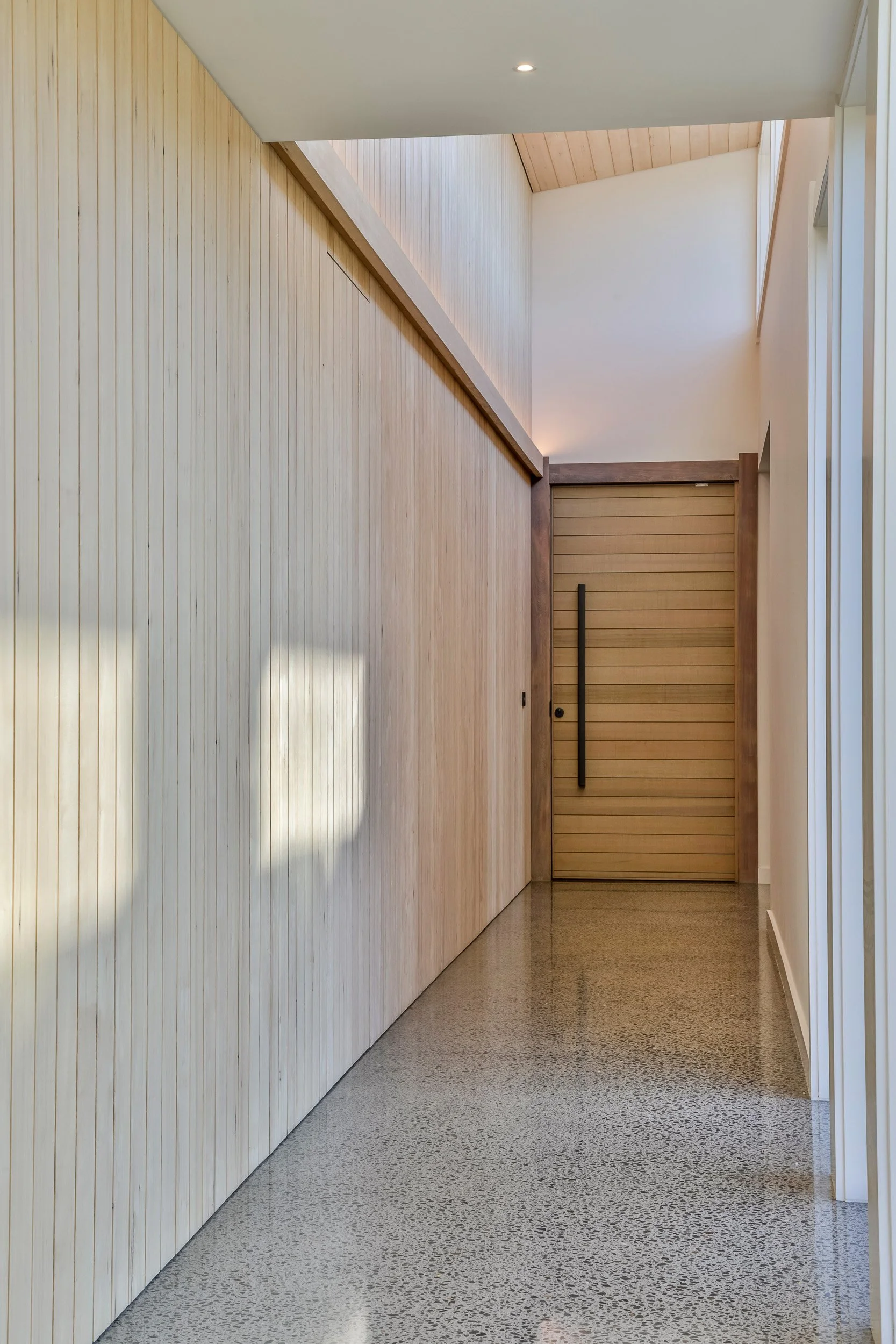 Minimalist hallway with light wood-paneled wall, polished concrete floor, and a wooden sliding door at the end. Taupo Architects. Home Design. Interior Design. New Zealand Architects. 