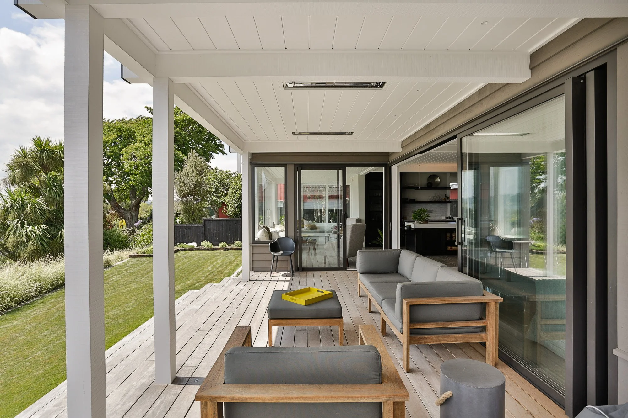 Modern covered patio with seating area and outdoor furniture, attached to a house with sliding glass doors, overlooking a landscaped backyard with trees and grass.