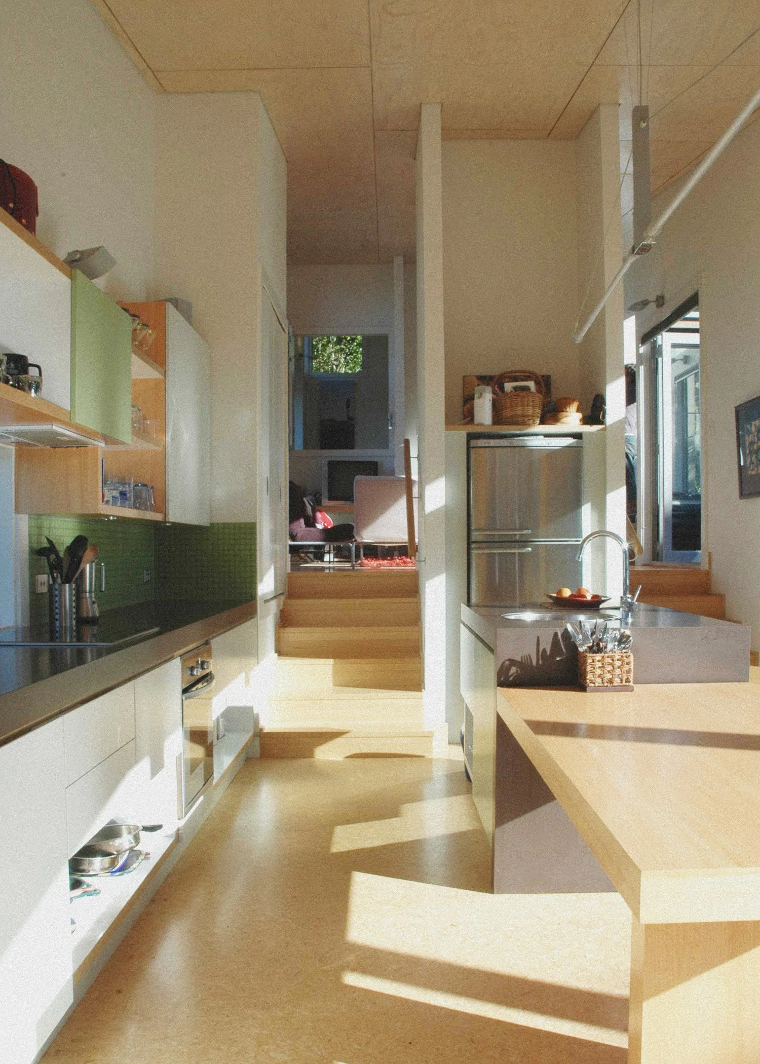Bright, modern kitchen with light wood cabinets and countertops, green tiled backsplash, open small island with a sink, and a view into a living room with chairs and a TV in the background. Bach Design New Zealand. Bach Architect. 