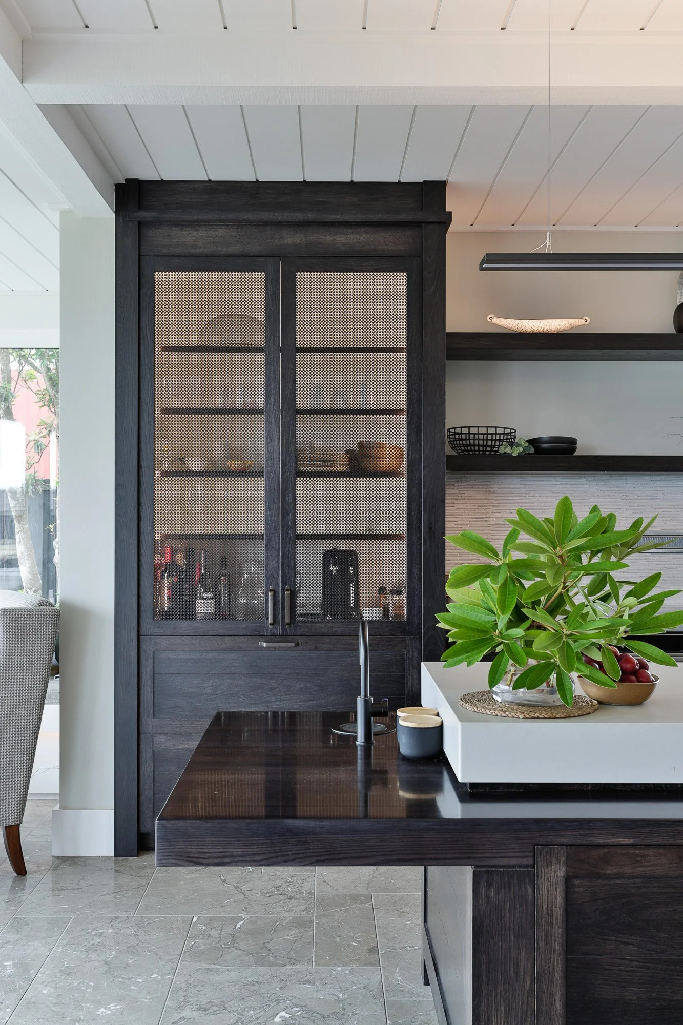 Interior of a modern kitchen with a black wooden pantry with mesh doors, a dark wood island with a sink, a white countertop with a plant and bowls, and open black shelves with black bowls and decorative objects.