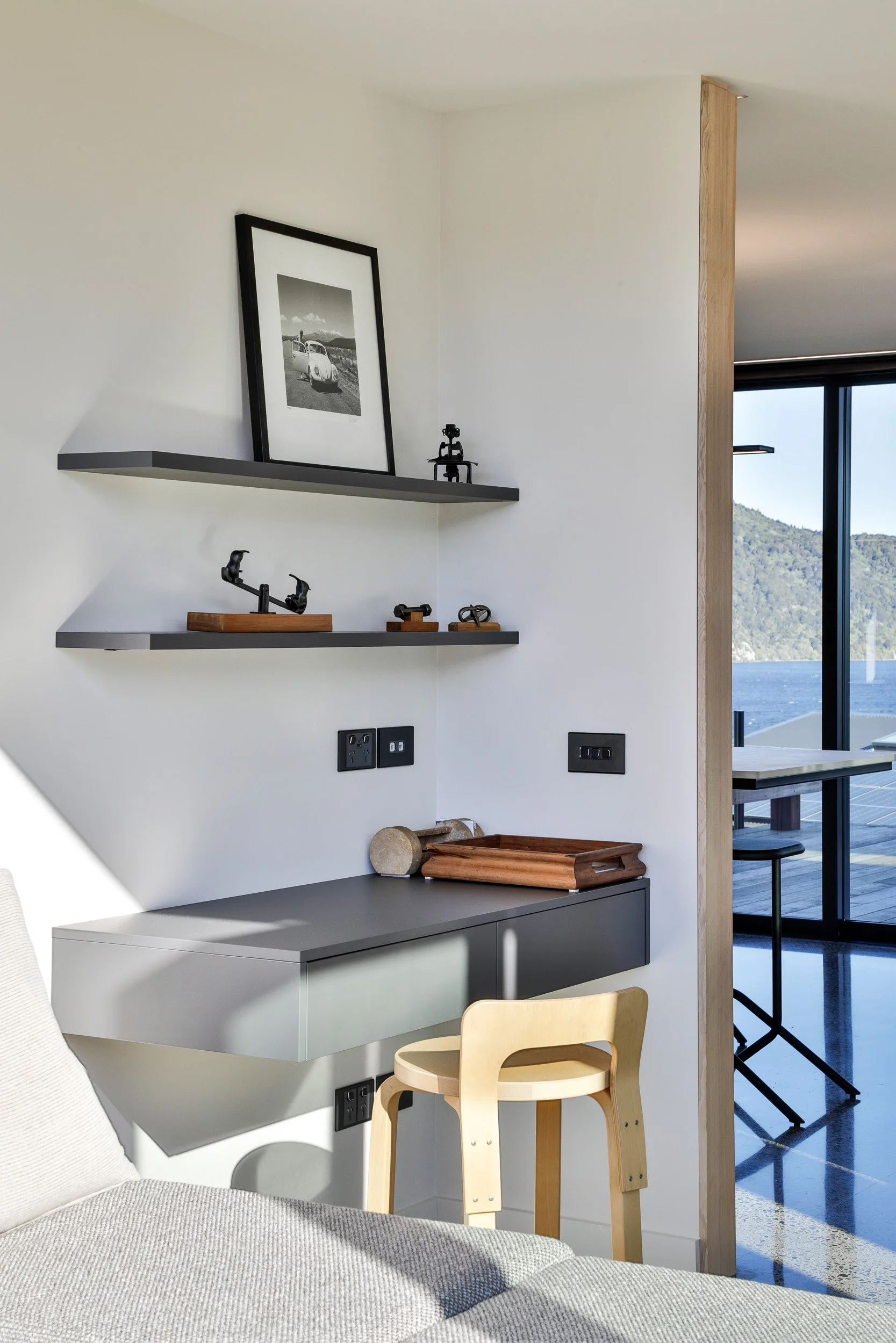Interior of a modern living space with floating shelves decorated with small sculptures and a framed black-and-white photo, a desk with a wooden tray, and a light wooden chair, overlooking a scenic view of water and mountains through large glass door