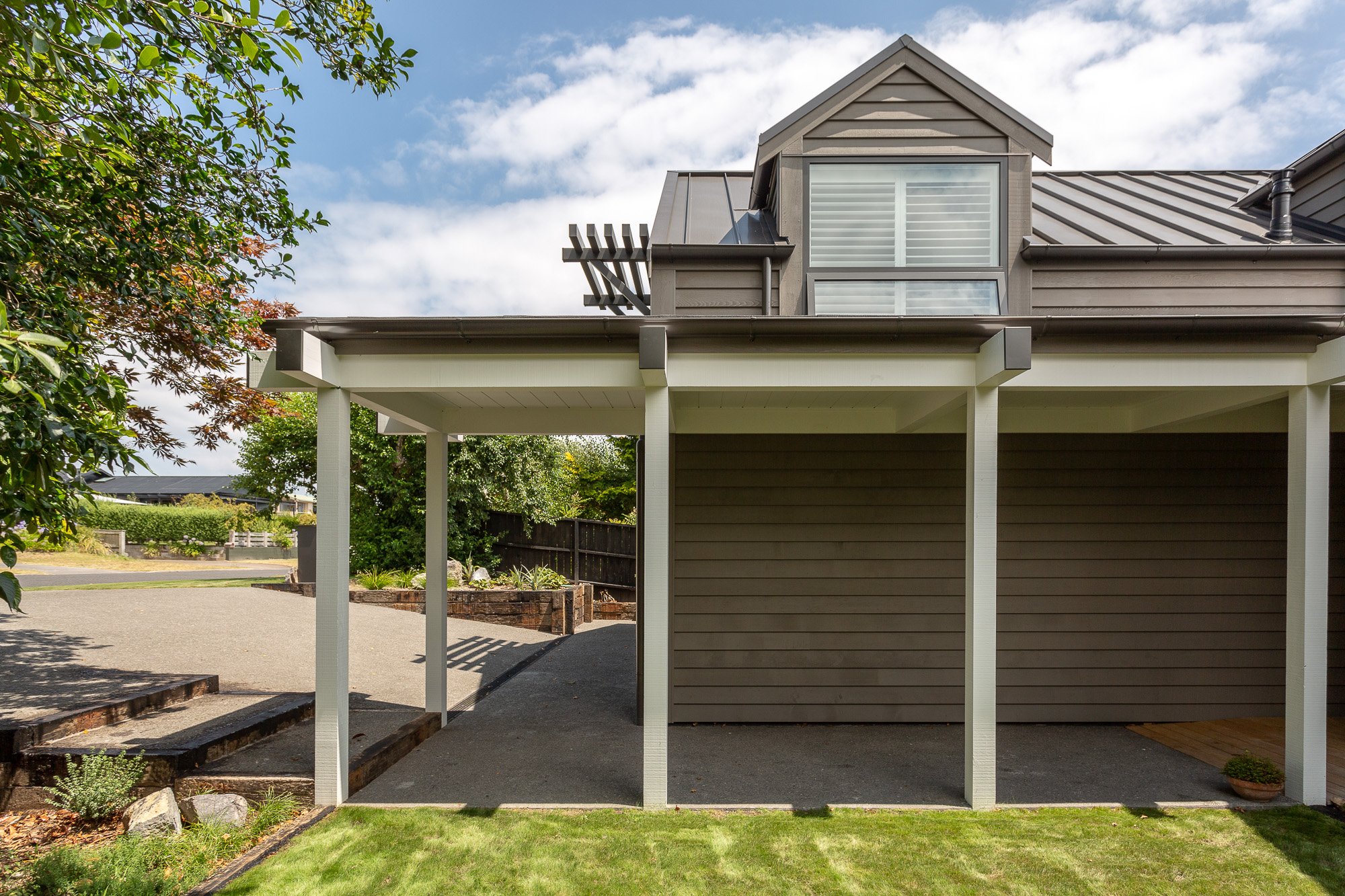 Rear view of a modern house with a covered concrete carport, gray wooden siding, a small upper dormer window, and a wooden deck on the second floor. Green lawn and landscaped garden in foreground, partly cloudy sky.