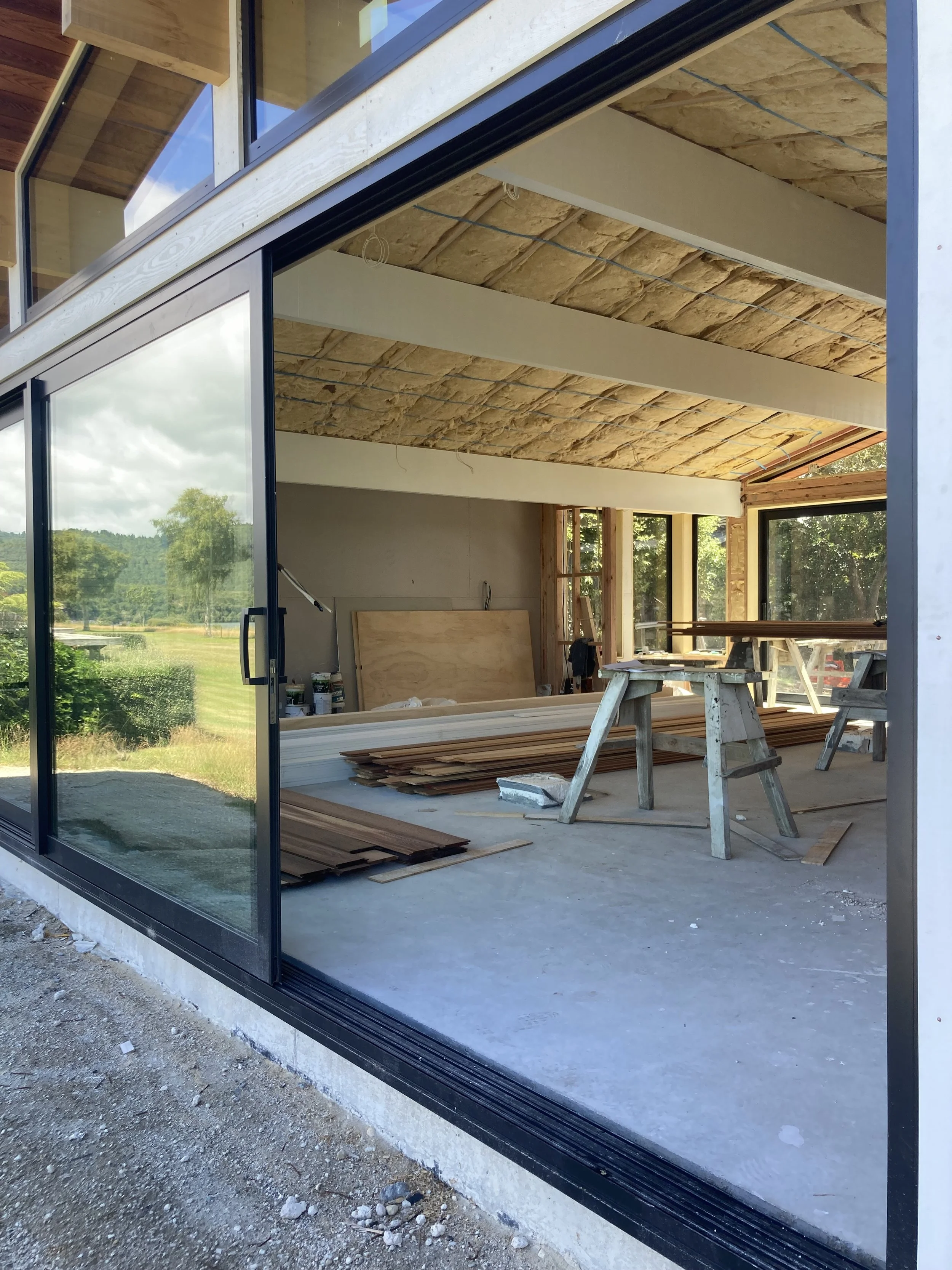 Interior of a house under construction, with wooden beams, construction materials, and tools inside, viewed through a large glass sliding door.