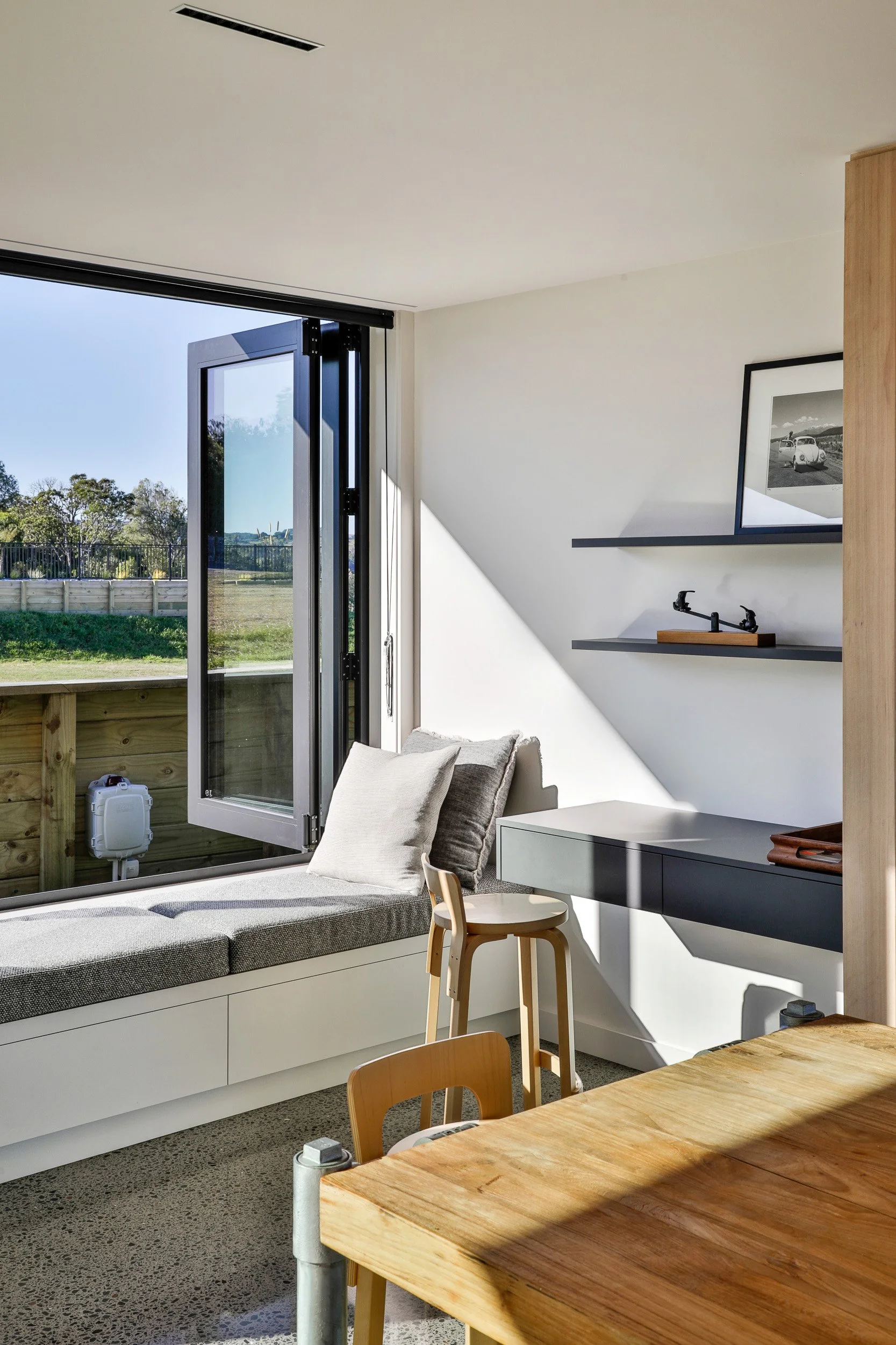 Sunlit modern living room with an open window, a cozy window seat with cushions, a small desk with a drawer, a framed picture of a car, and a wooden table with chairs. Taupo Architects. Home Design. Interior Design. New Zealand Architects. 