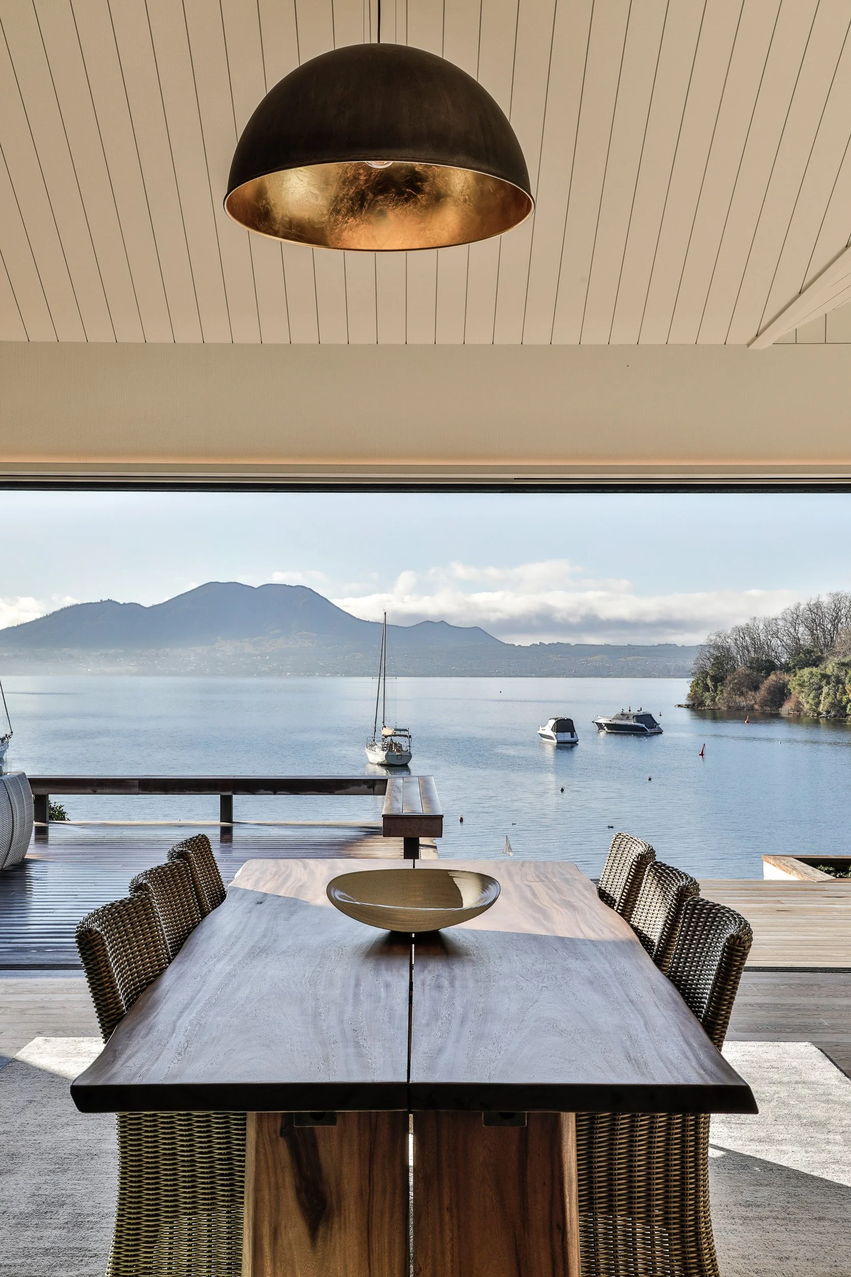 A dining room with a view of Lake Taupo and Mt Tauhara, featuring a wooden dining table, six chairs, a bowl on the table, and a black pendant light hanging from the ceiling. Taupo Architects. The Craftsmen Taupo Builders. 