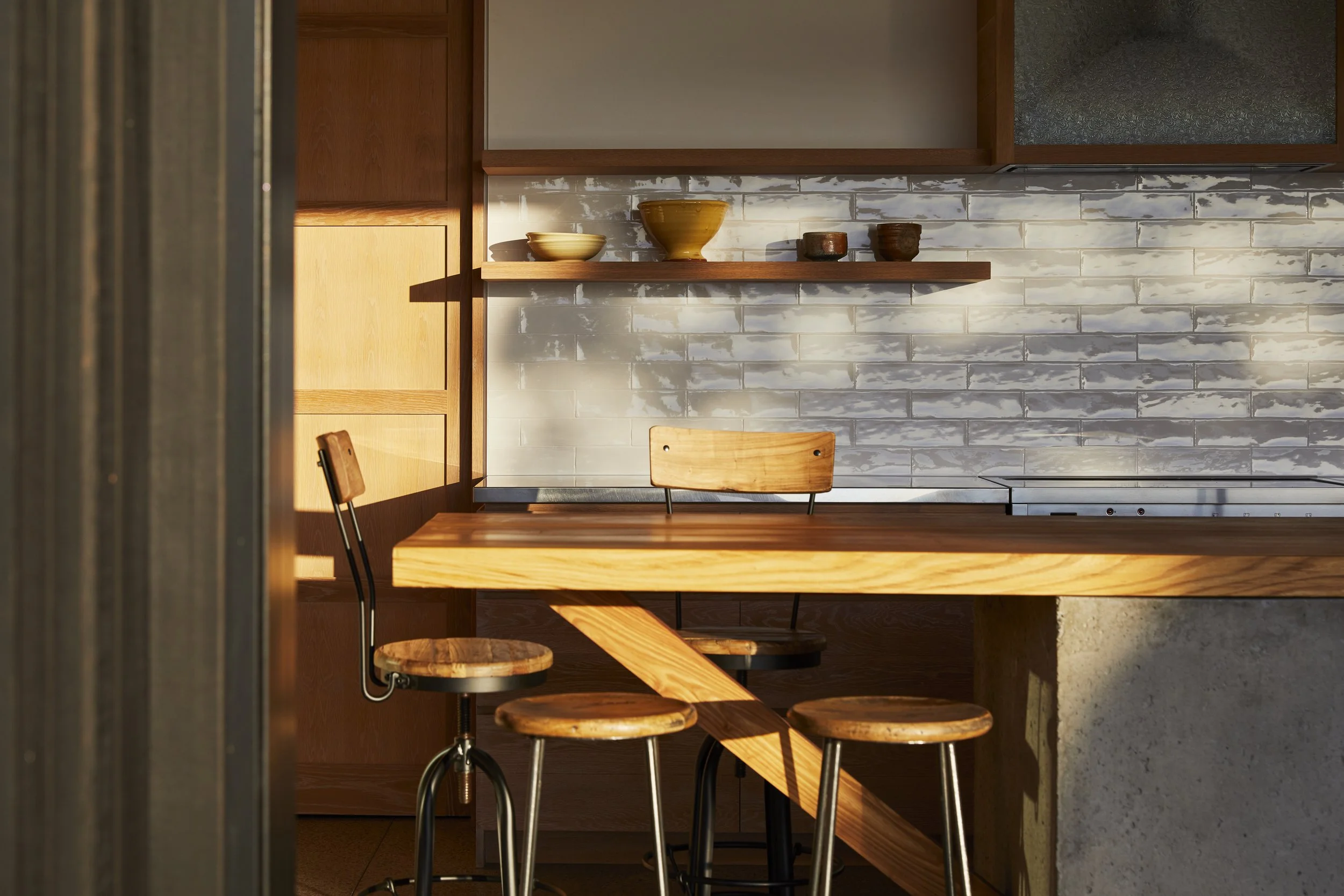 Sunlit kitchen with a wooden breakfast bar, four matching stools, a wooden chair, open shelving, a white tile backsplash, and ceramic bowls and cups.