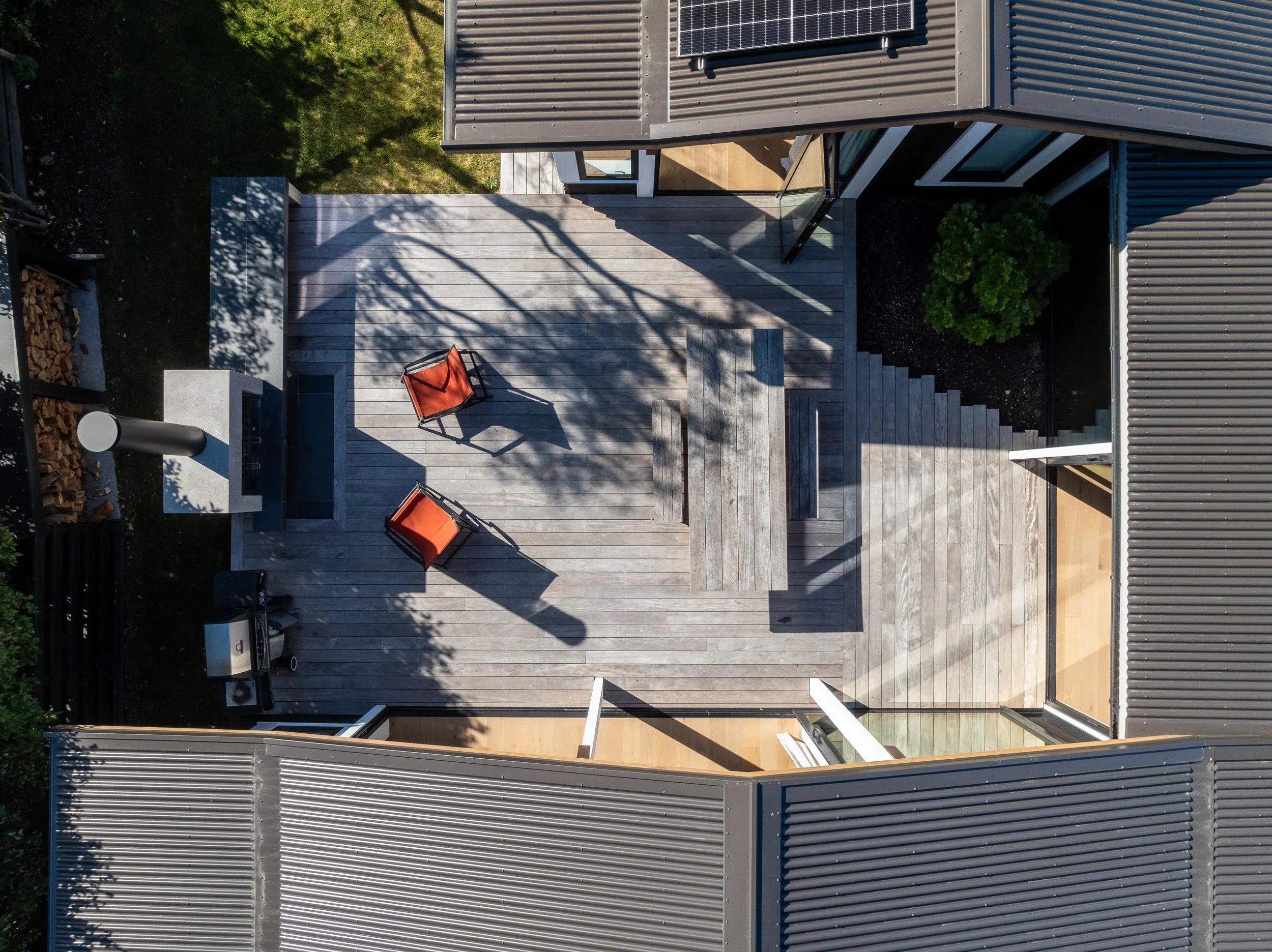 Aerial view of a modern house's outdoor patio with two red chairs, a wooden table, steps, and a barbecue grill, surrounded by greenery and metal siding roofs.