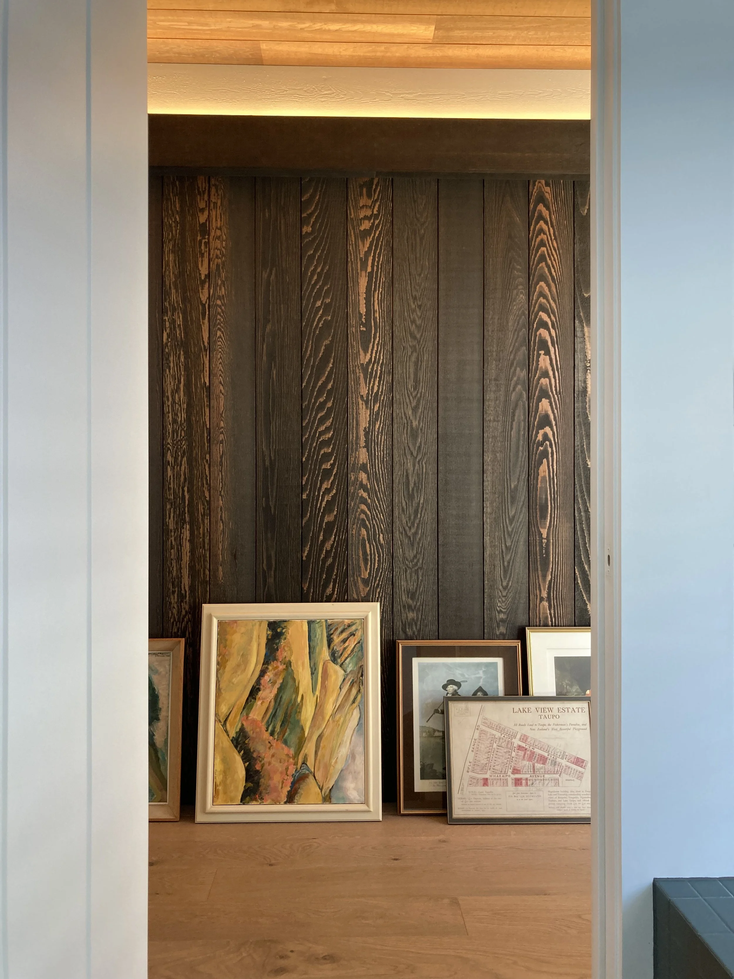 Interior view of a room with artwork and framed photos propped against a dark wooden wall, with a lighter wooden floor and ceiling.