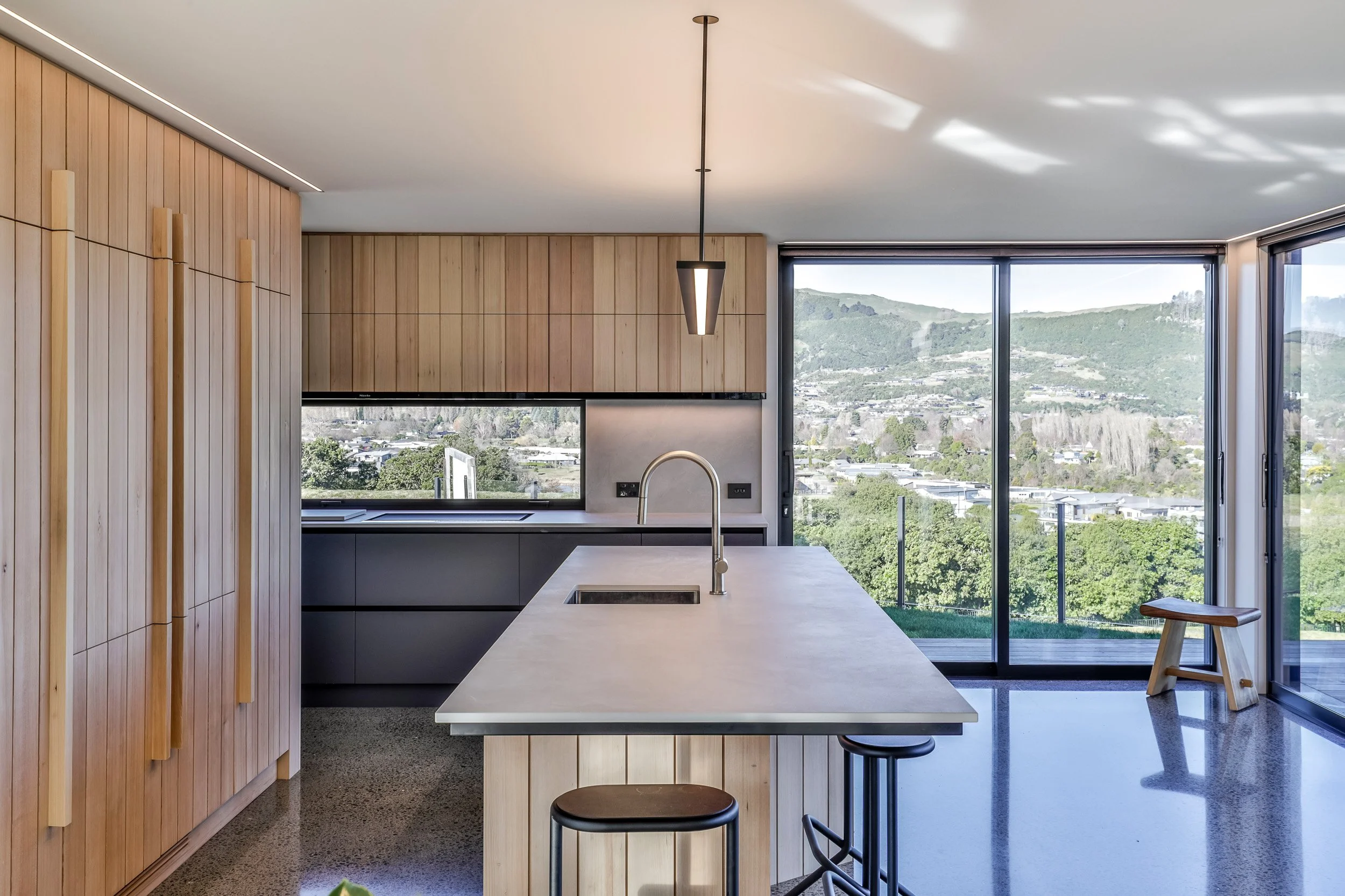 Modern kitchen with wooden cabinets, white marble island, black countertops, large glass sliding doors, and views of greenery and hills outside. Taupo Architects. Home Design. Interior Design. New Zealand Architects. Kitchen Design Taupo. 