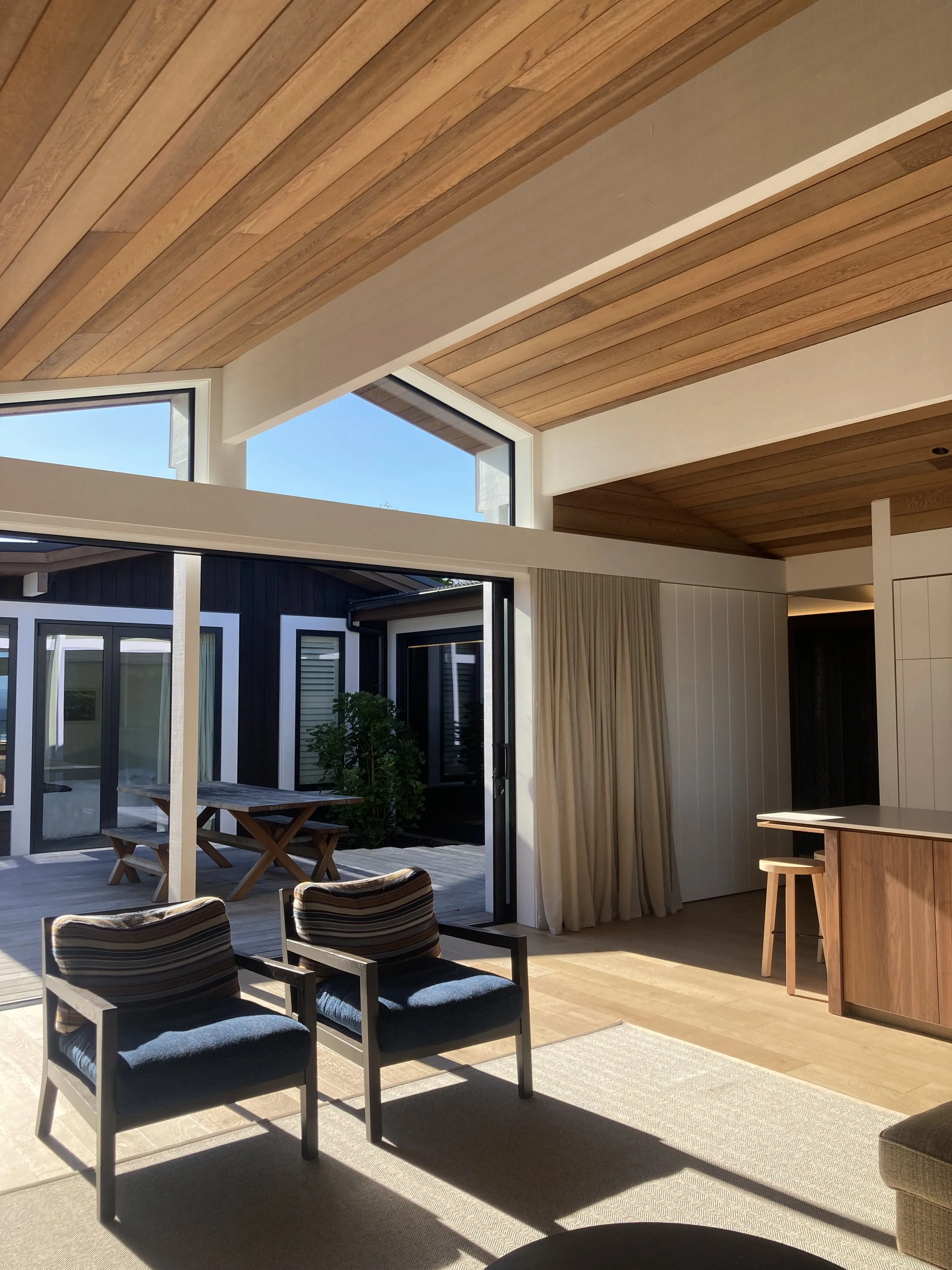 Living room with wooden ceiling, large windows, and french doors leading to an outdoor patio with a picnic table. Two striped armchairs and a rug are in the foreground.