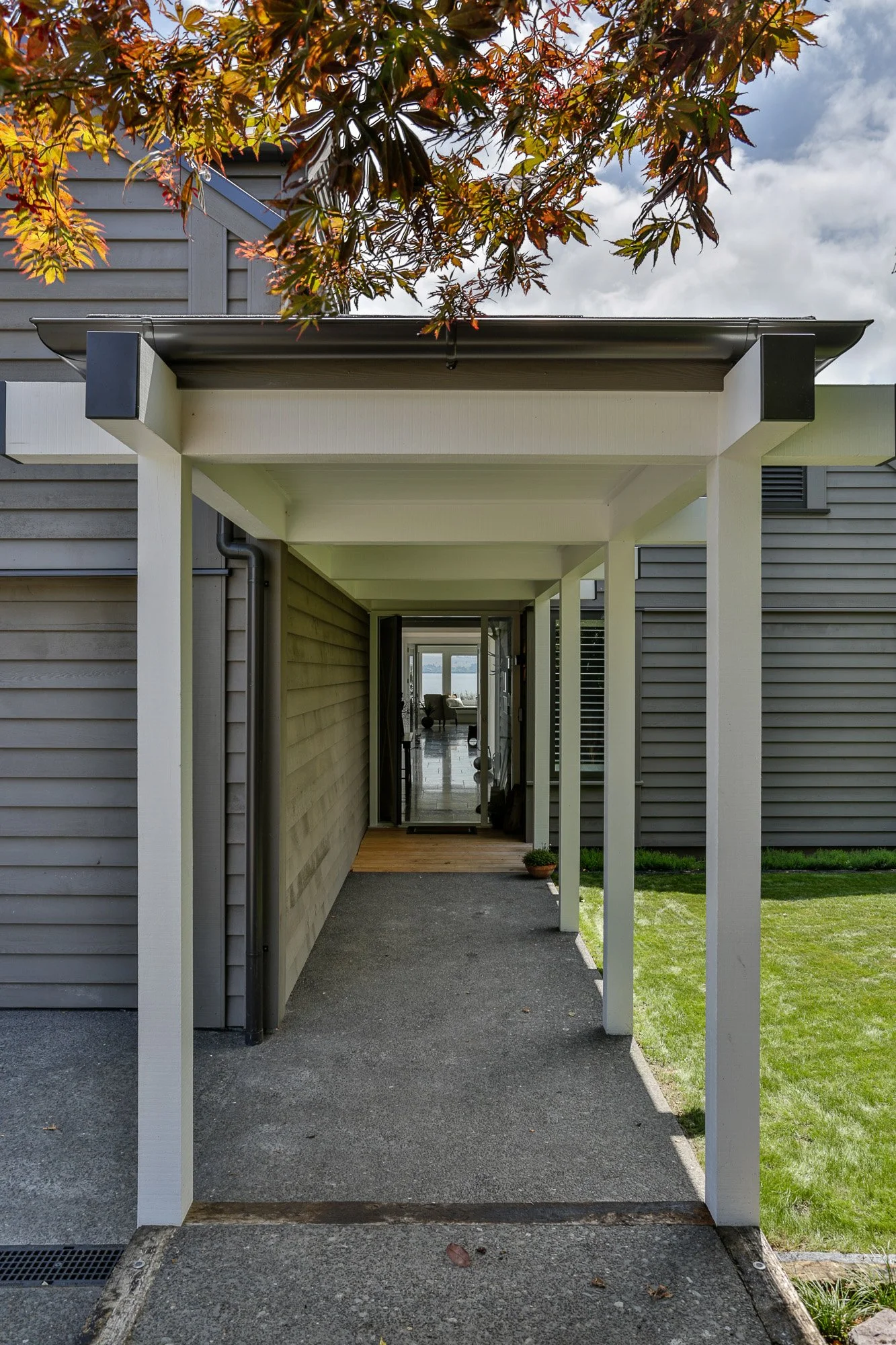 View of a covered walkway leading to a house, with a grassy lawn on the right and a tree with red and green leaves above.