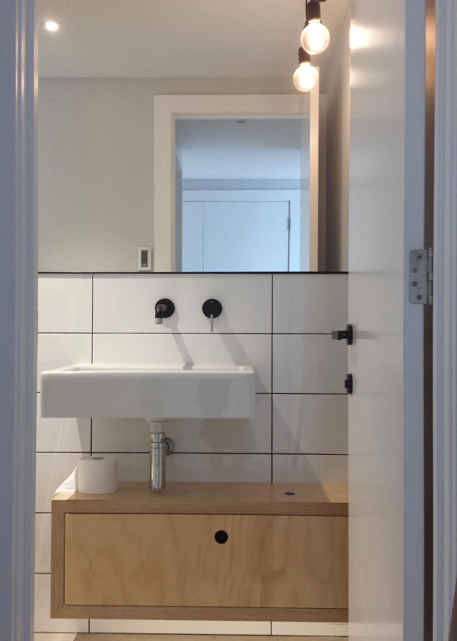 Minimalist bathroom with white tiled wall, black fixtures, a small white sink, a mirror above, and a wooden cabinet below, with exposed light bulbs overhead. Bathroom design Taupo. Interior design Taupo. Haimes Builders Taupo. 