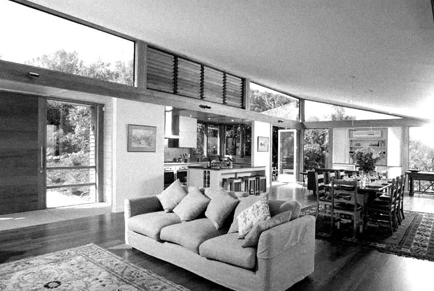 Living room with a sofa and cushions, dining table with chairs, large windows, and an open kitchen area.