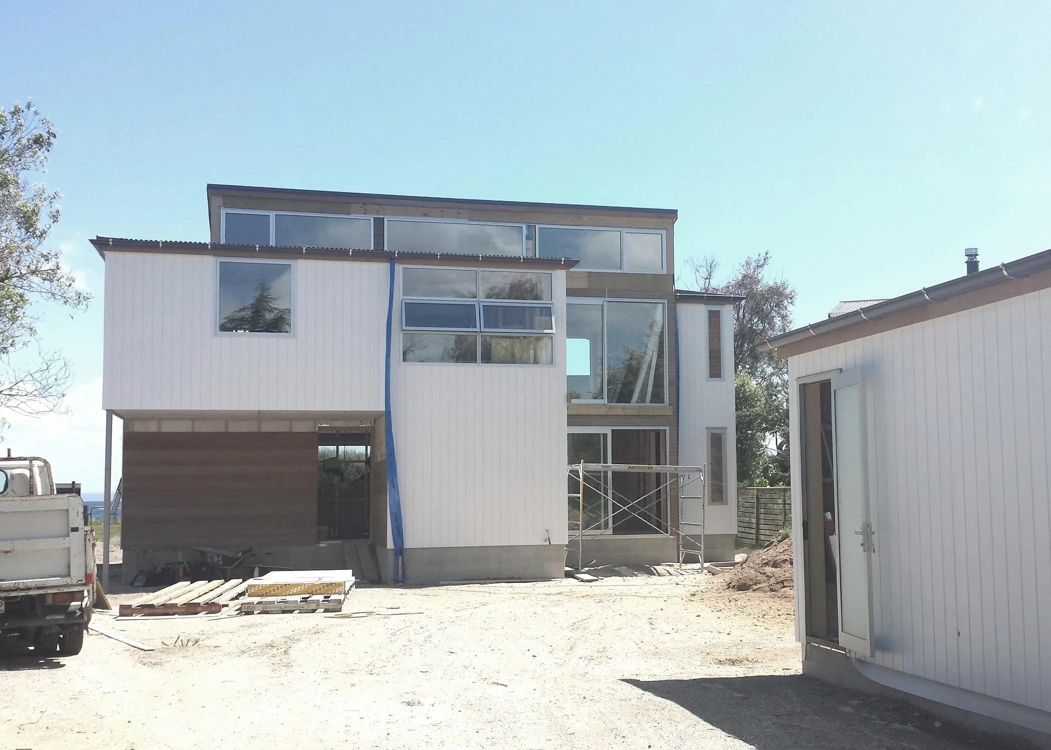 Under construction modern multi-story house with large windows and scaffolding, surrounded by construction materials and vehicles. Bach Design New Zealand. Bach Architect. Holiday Home Architect. Rural Architect. 