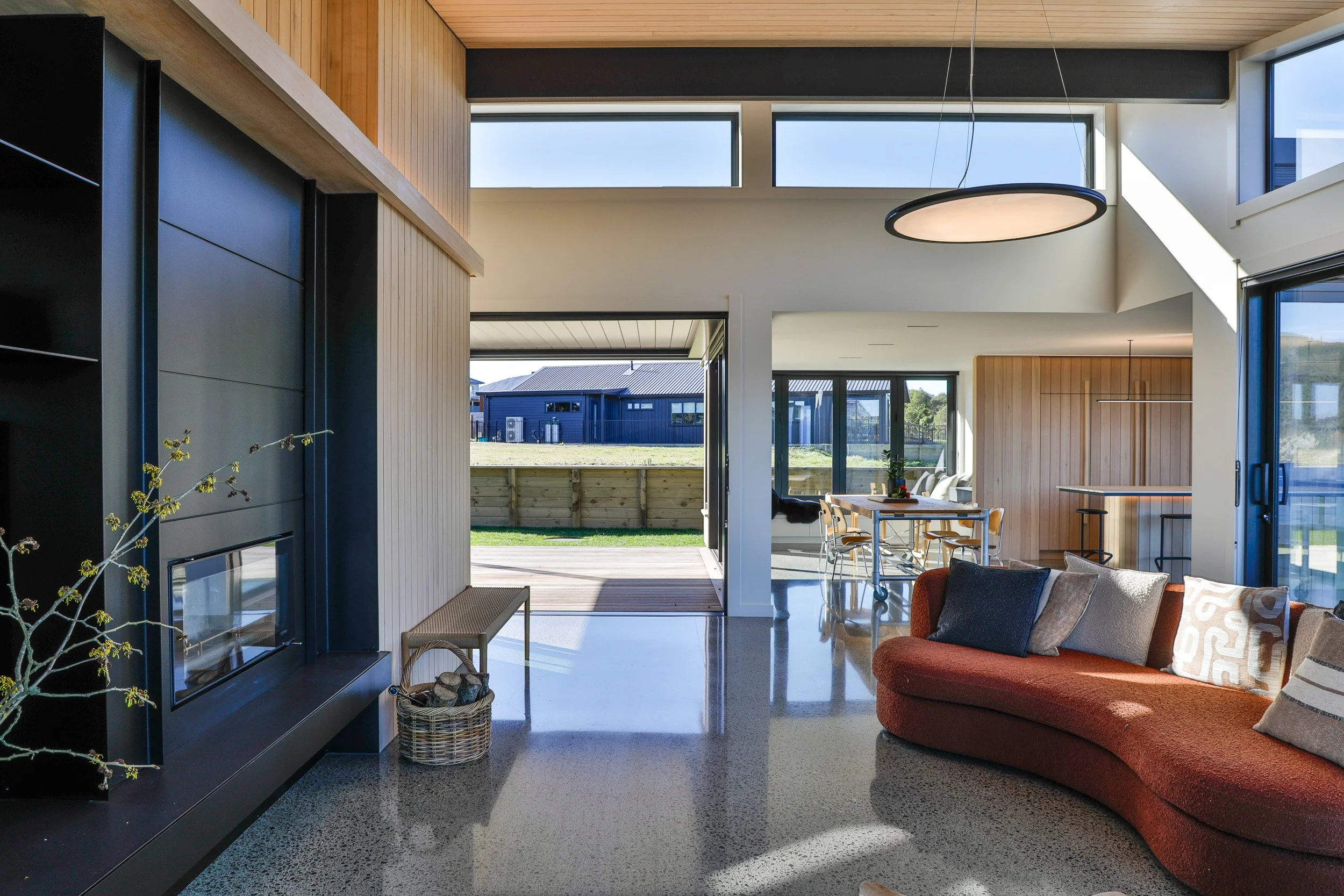 Modern living room with large windows, wooden accents, a curved orange sofa with pillows, a dining table with chairs, and an open door leading to a backyard. Taupo Architects. Home Design. Interior Design. New Zealand Architects. 