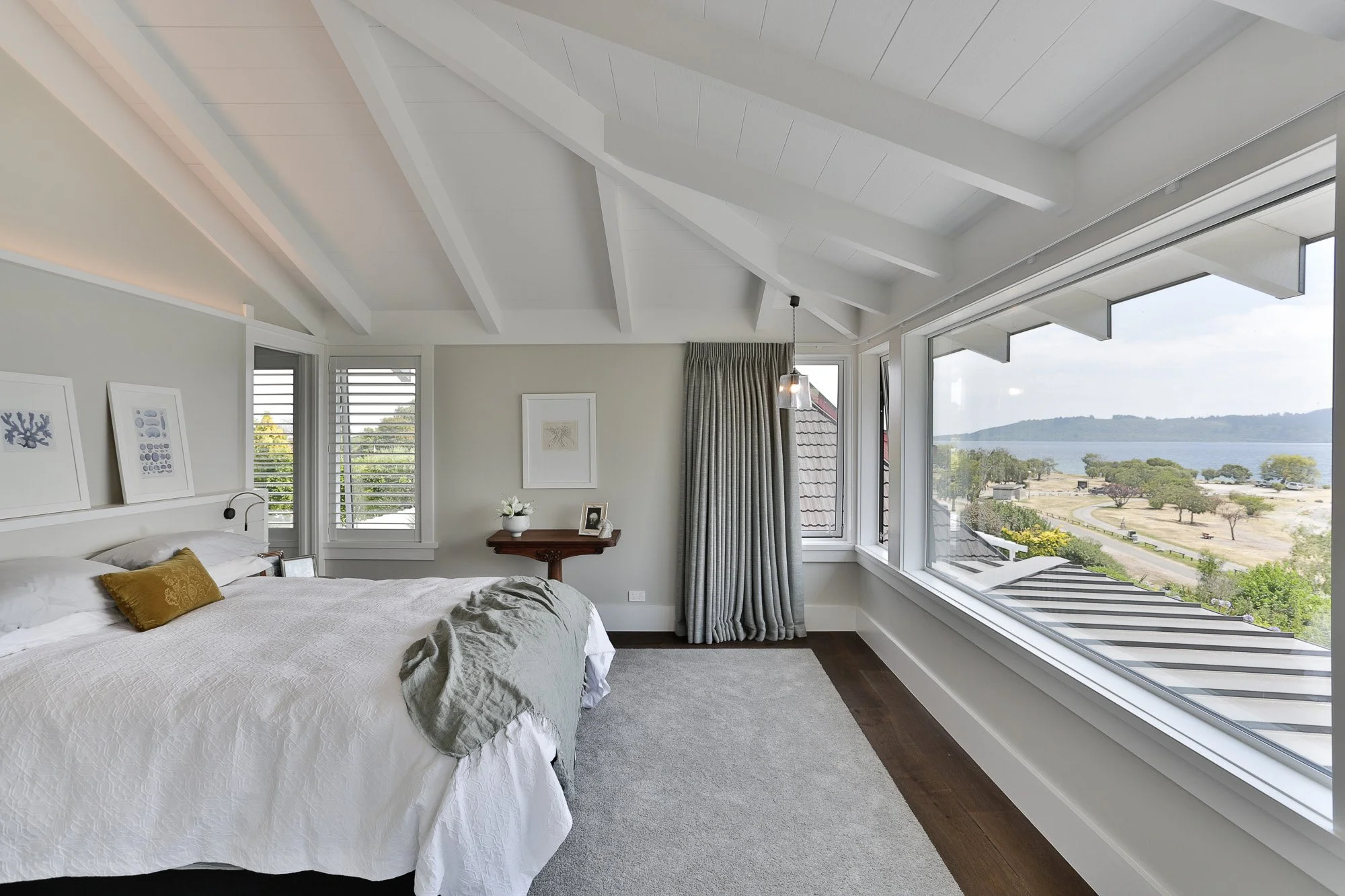 Bright bedroom with large window view of water and landscape, white walls, vaulted ceiling with exposed beams, bed with white bedding and a small yellow pillow, grey curtains, and wooden furniture.