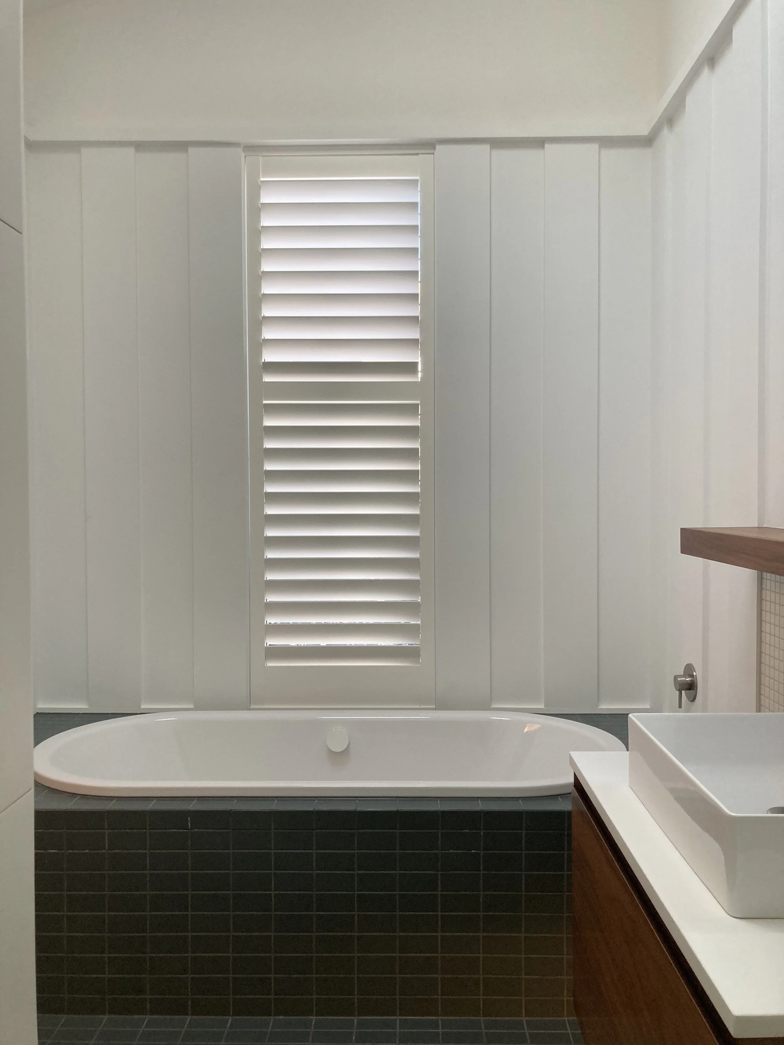 Modern bathroom with a white bathtub, dark tiled exterior, white paneled walls, and a window with white shutters.