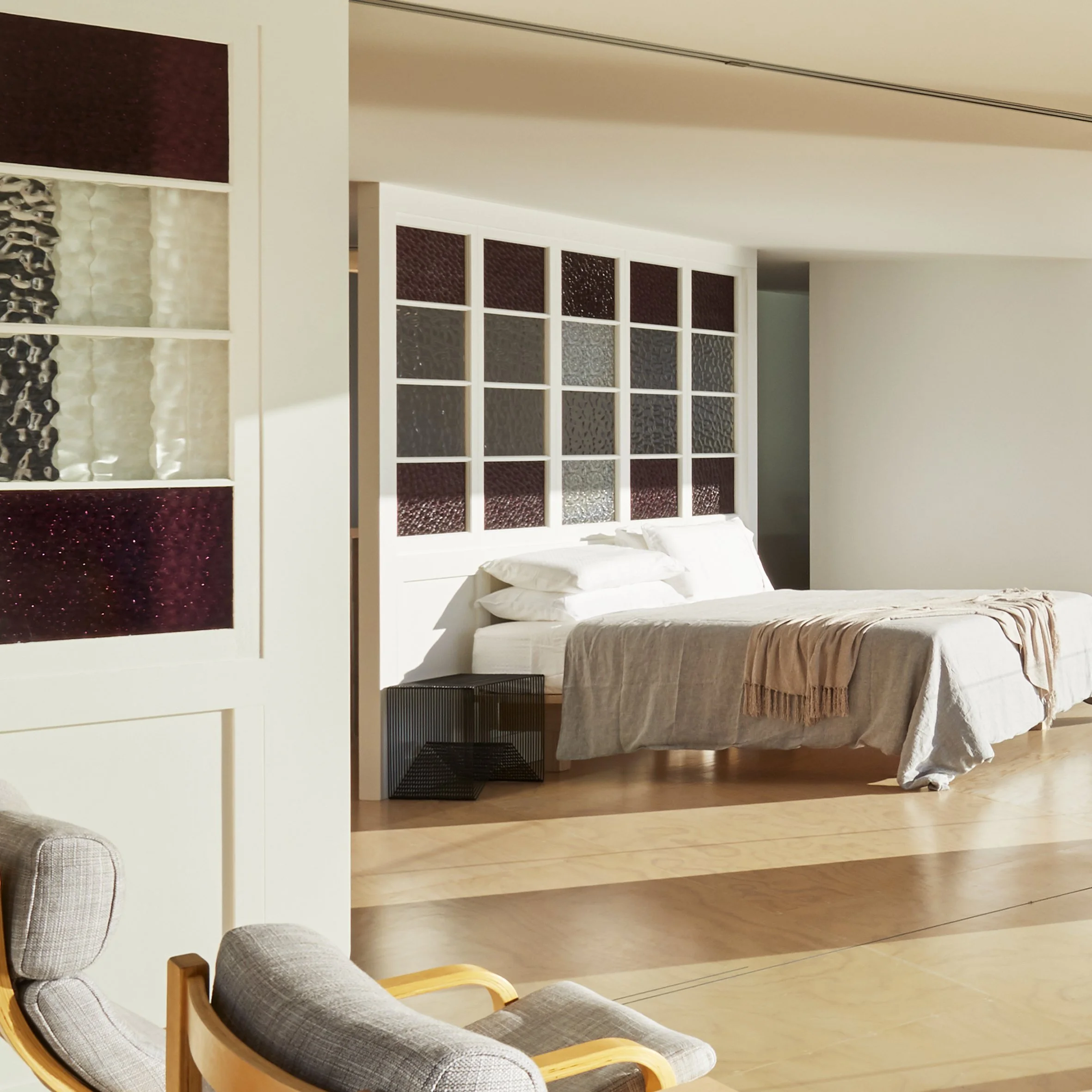 A bedroom with a bed, pillows, and a beige blanket, surrounded by white walls and stained glass accents in the headboard and partition, with two gray armchairs in the foreground.