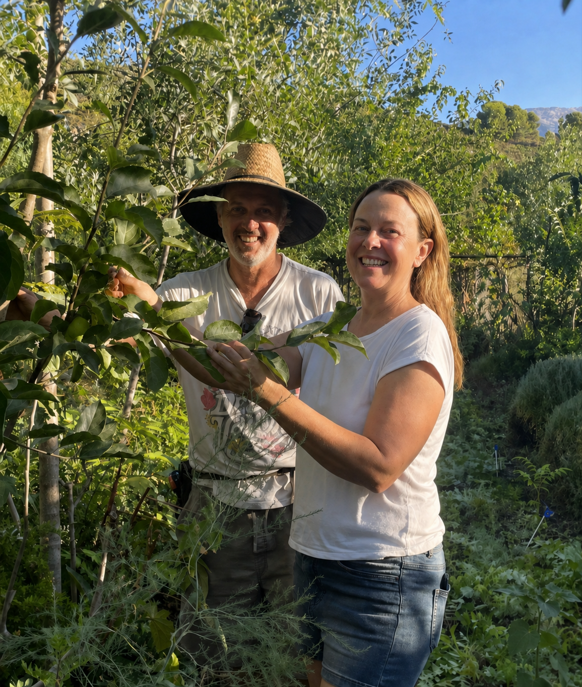 A man wearing a wide-brimmed straw hat and a woman with curly hair smiling and holding a branch with green leaves in a lush garden.